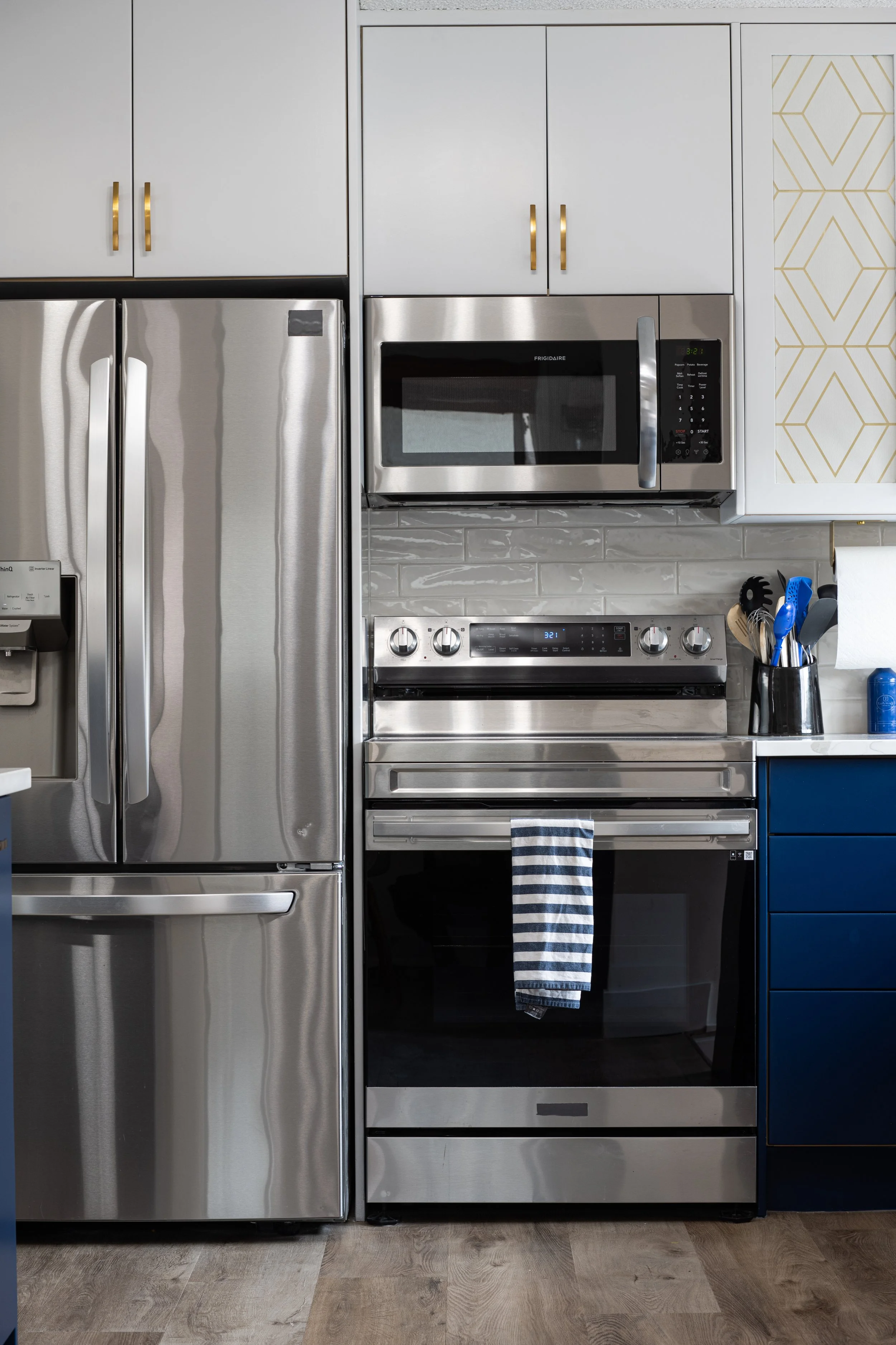 stainless steel appliances are shown in a scene of white upper cabinets with blue lower cabinets.  Gold handles are featured as well as a gold pattern wallpaper in the glass of a glass door.  The backsplash is a sleek wavy subway tile pattern.