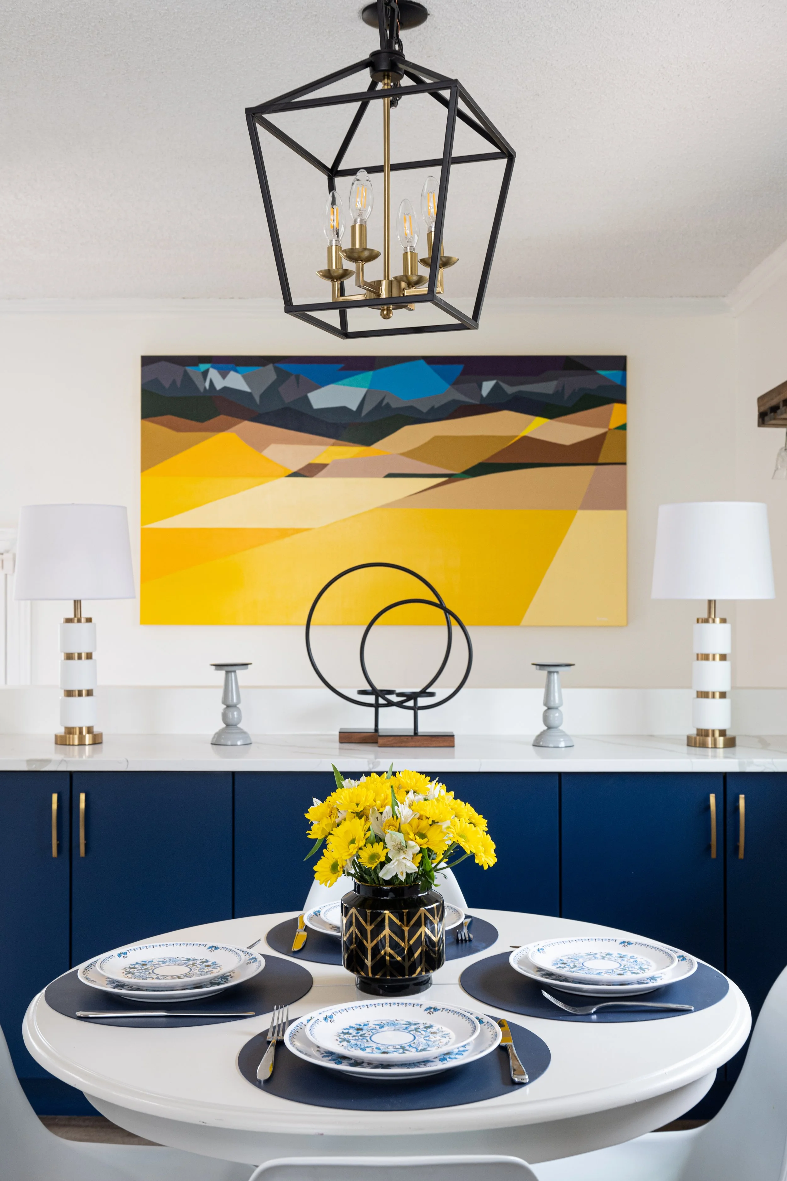 a freshly renovated dining room is nicely styled.  The background shows a set of lower cabinetry painted blue, and a nice piece of yellow and blue complimentary artwork is shown above it.  A grey table with blue accents, and bright fresh flowers