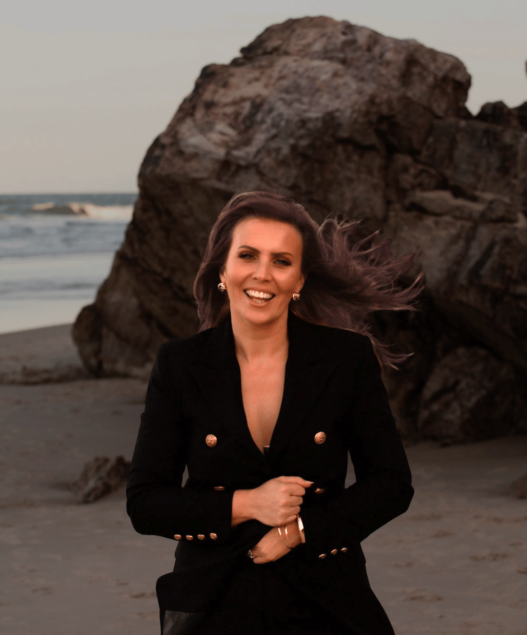A woman smiling and running on a beach with large rocks in the background at sunset.
