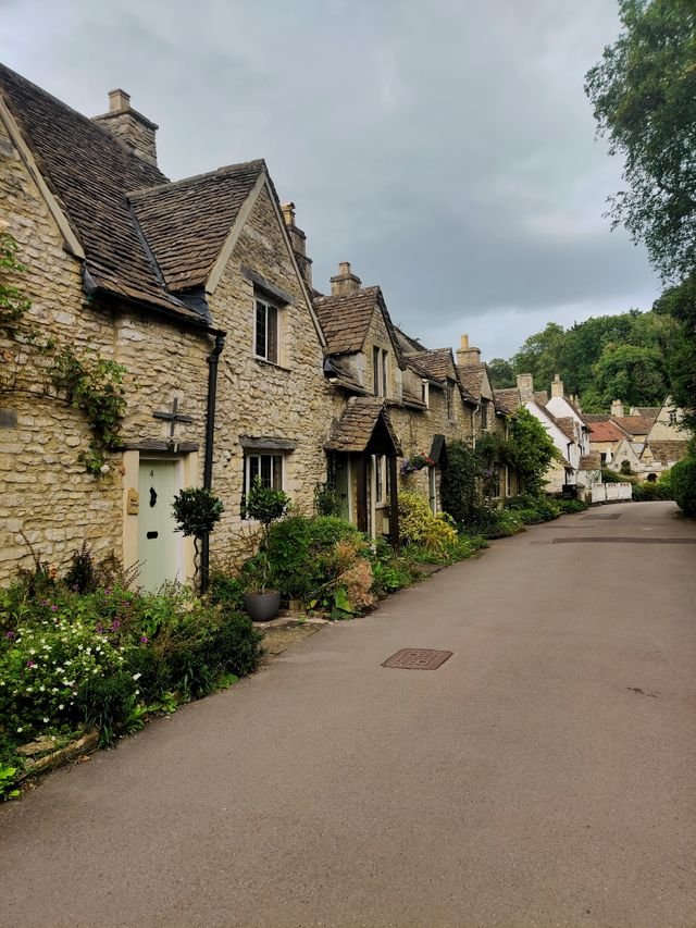 Historic stone cottages with tiled roofs and small front gardens along a narrow curved street in a rural village under cloudy sky.