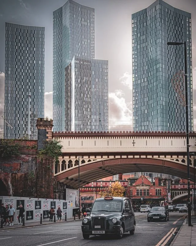 A view of city skyscrapers across a red and cream coloured iron rail bridge in Manchester.