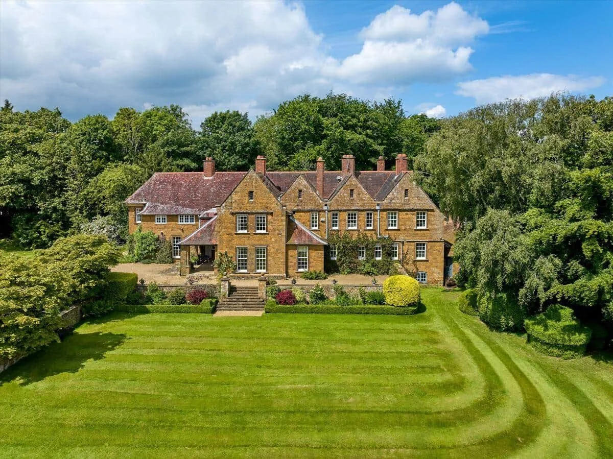 A large stone manor house with chimneys in front of an expansive mowed lawn.