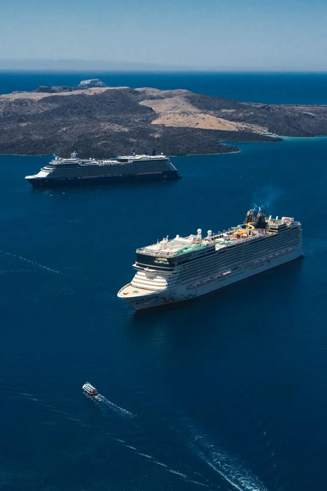 Two white cruise ships sailing on the dark blue ocean past a remote island coastline.