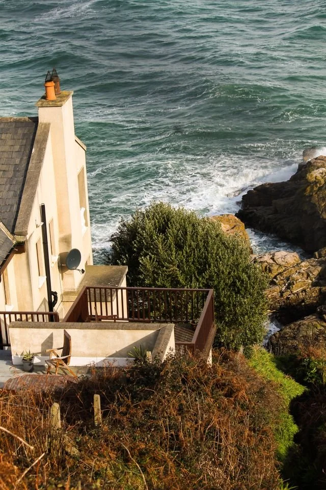 A white house with slate roof on a cliff with trees, overlooking the sea.
