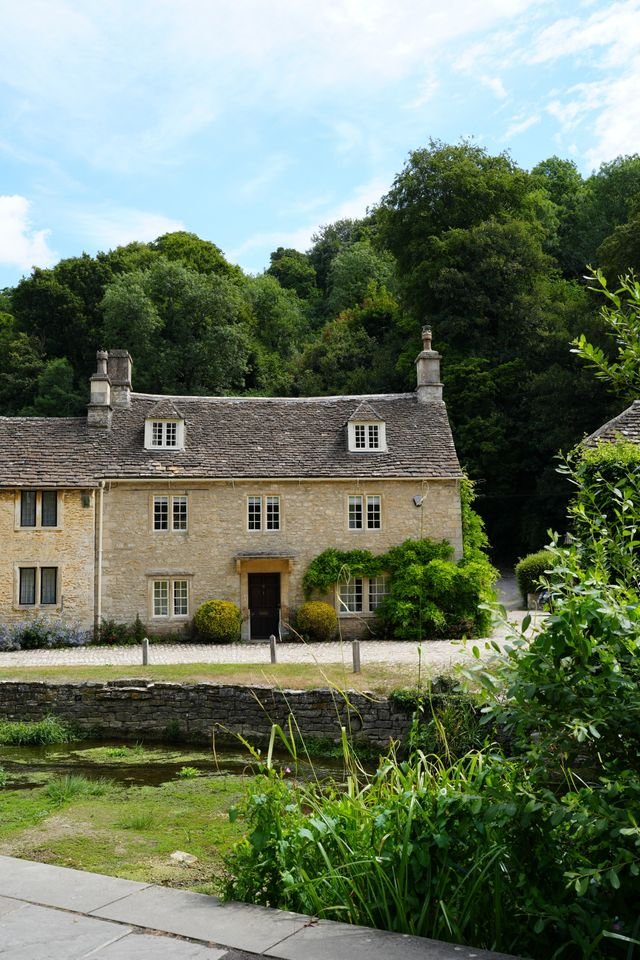 An old stone cottage with slate roof in front of green trees and blue sky.