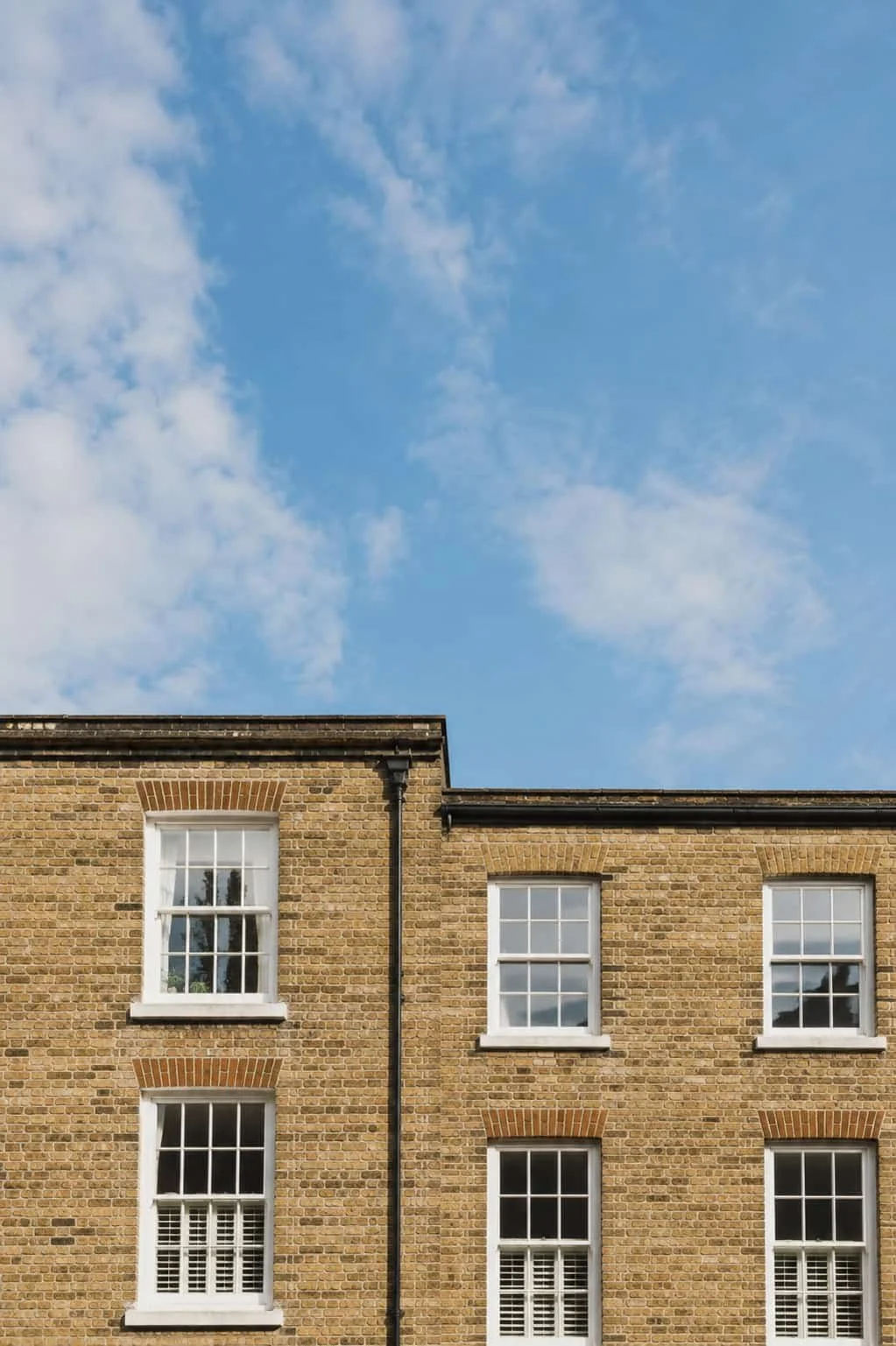 Part of a brick building with four white-framed windows and a black drainpipe, set against a bright blue sky with scattered clouds.