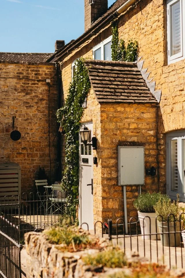 A quaint yellow brick terraced house with white window shutters and ivy climbing the walls.