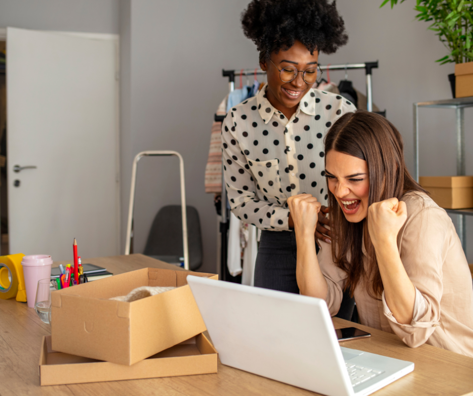 Two women celebrating at a desk with a laptop in an office or retail space, one sitting and showing excitement, the other standing and smiling.