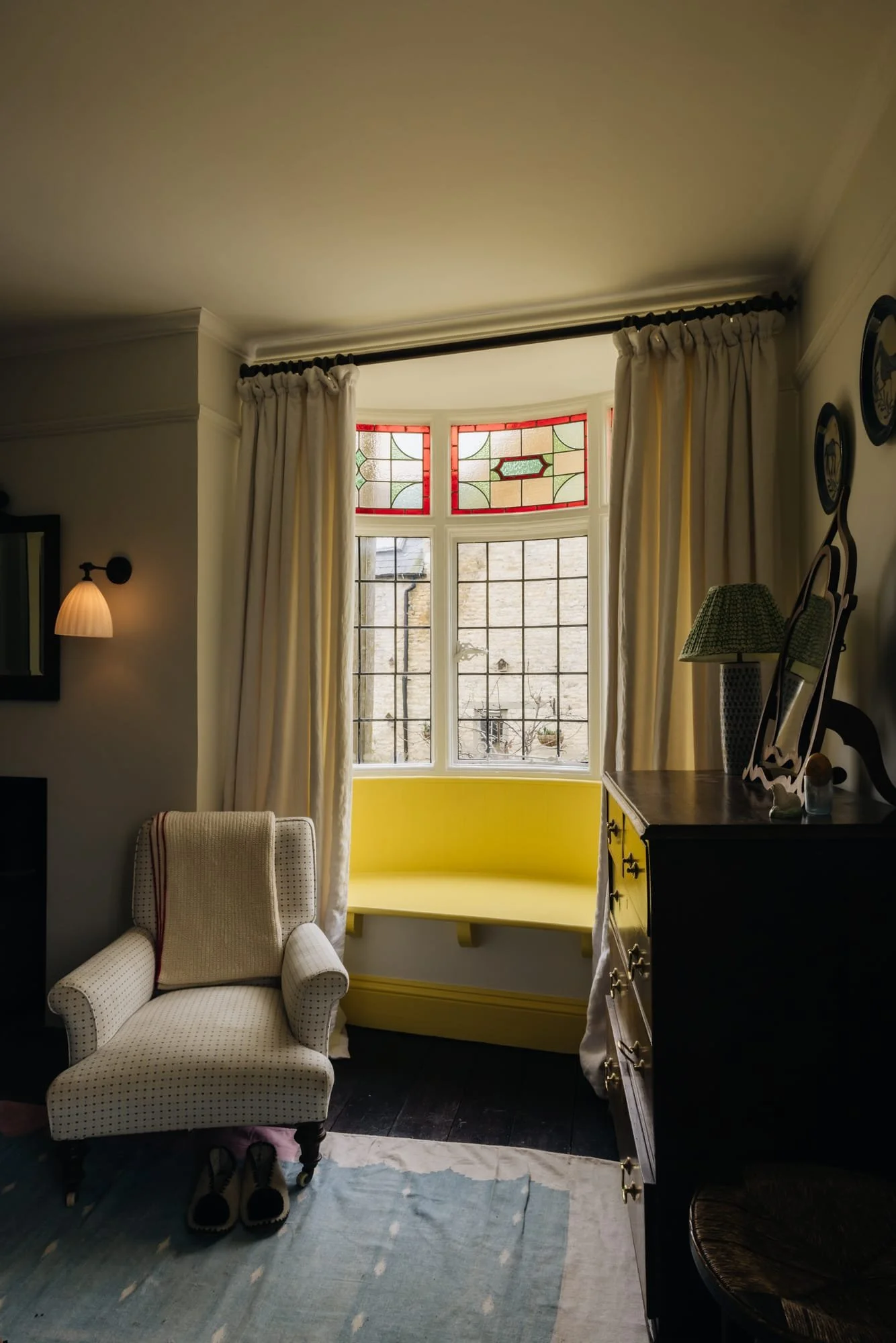 A cozy living room featuring a white armchair, a dark wooden dresser with a mirror, a table lamp, and a large bay window with stained glass at the top, surrounded by cream curtains, with a light blue rug on dark hardwood floors.