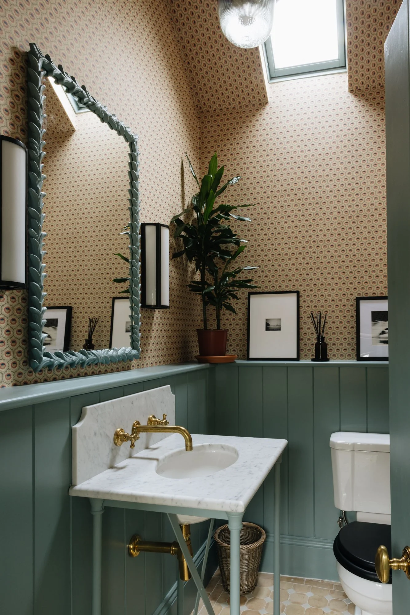 A small vintage-style bathroom with patterned wallpaper, green wainscoting, a marble-topped sink with gold fixtures, a mirror with a decorative frame, black and white photographs, a potted plant, framed pictures, reed diffusers, a laundry basket, and