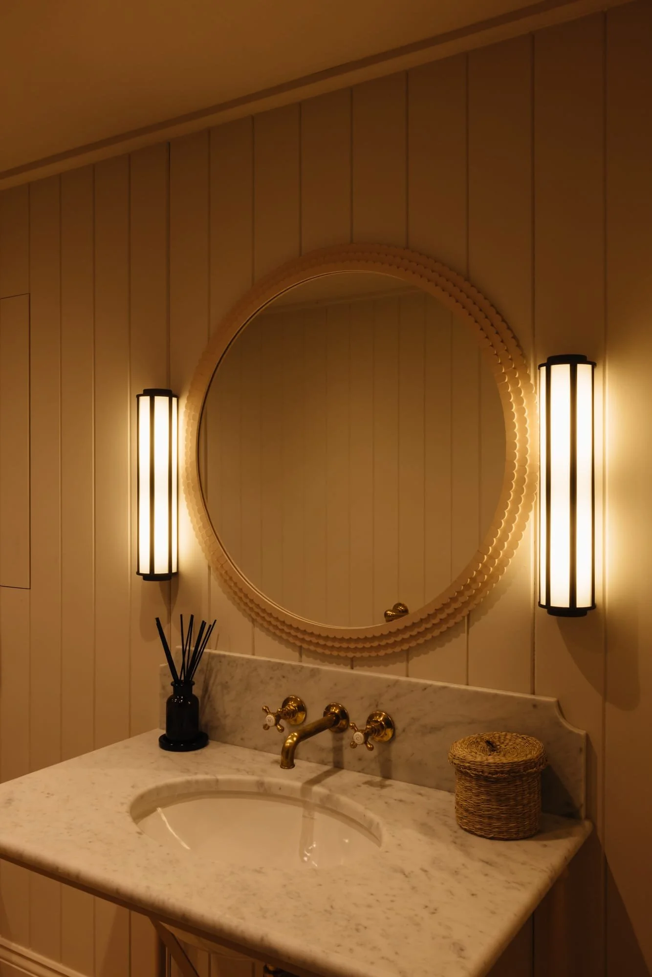A bathroom vanity with a marble countertop, a round mirror with a decorative frame, two vertical wall sconces emitting warm light, a black diffuser with reed sticks, and a small woven basket.