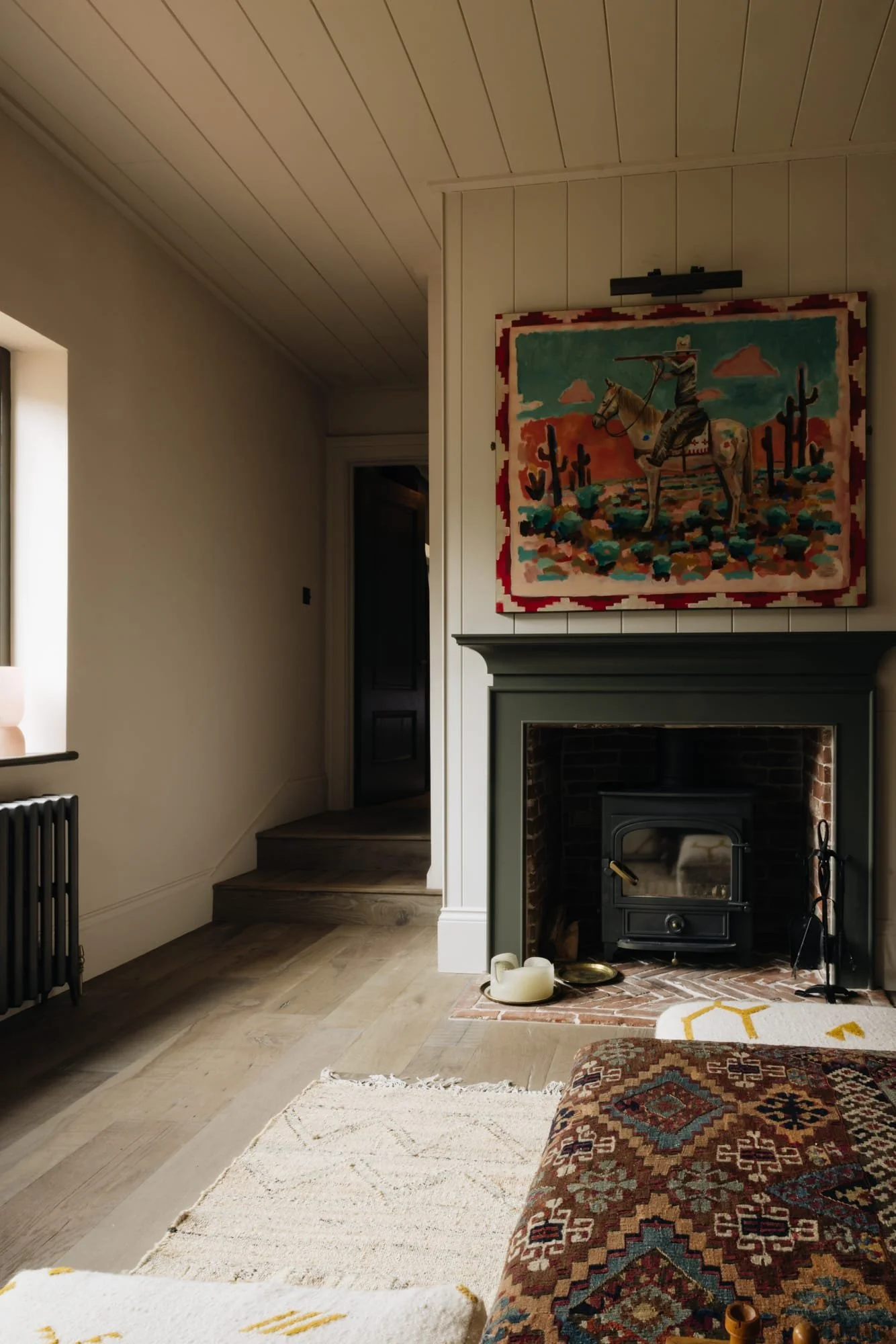 Living room with a fireplace topped by a colorful painting of a cowboy on a horse and cacti in a desert landscape. The room has a wooden ceiling, and a window on the left allows natural light. Part of a patterned sofa and a white rug are visible.