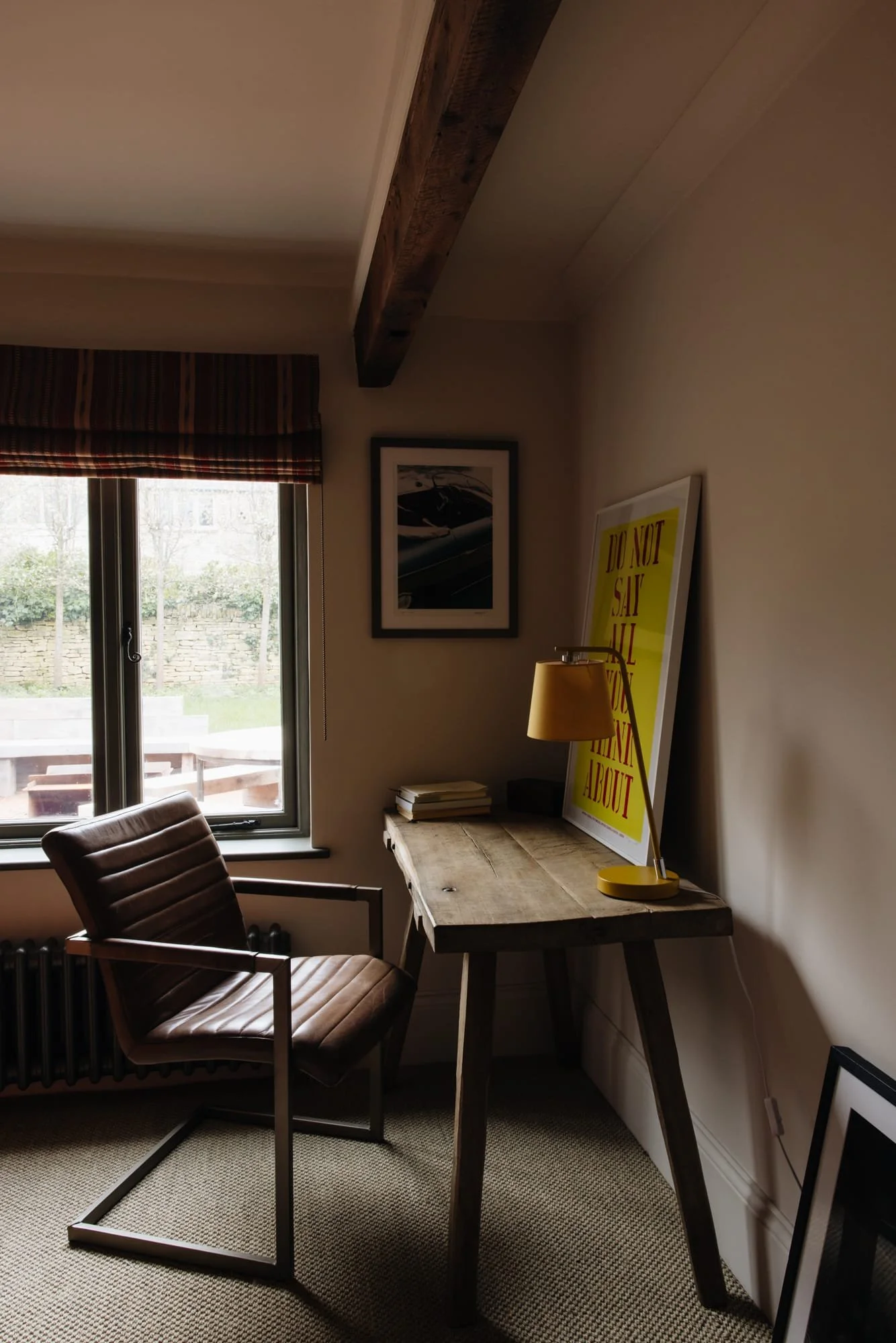 A cozy corner of a room with a window, a brown leather chair, and a rustic wooden desk with a yellow table lamp and framed artwork.