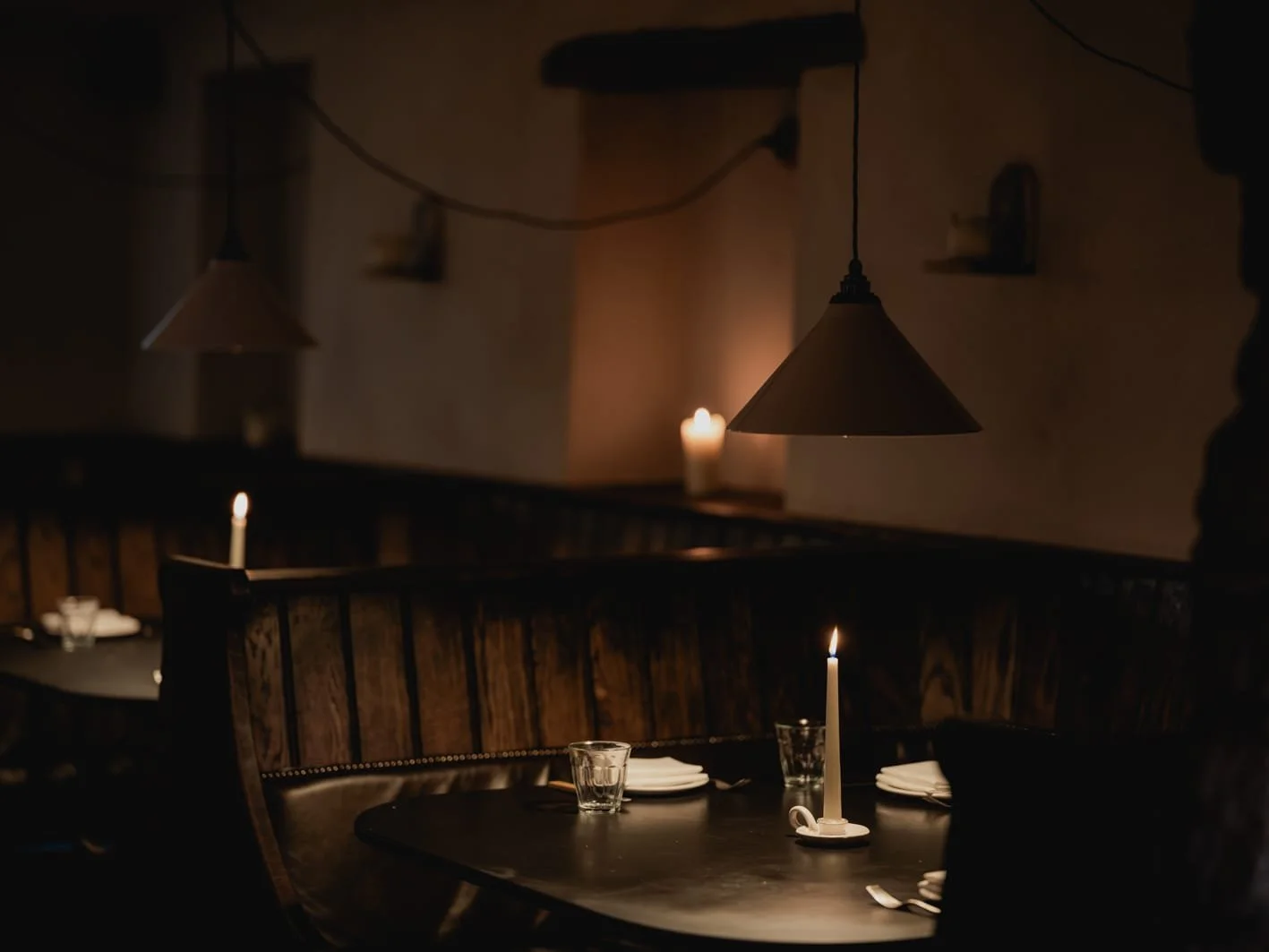 A dimly lit restaurant interior with candles on tables, hanging pendant lights, and minimal decor, creating a cozy and intimate atmosphere.