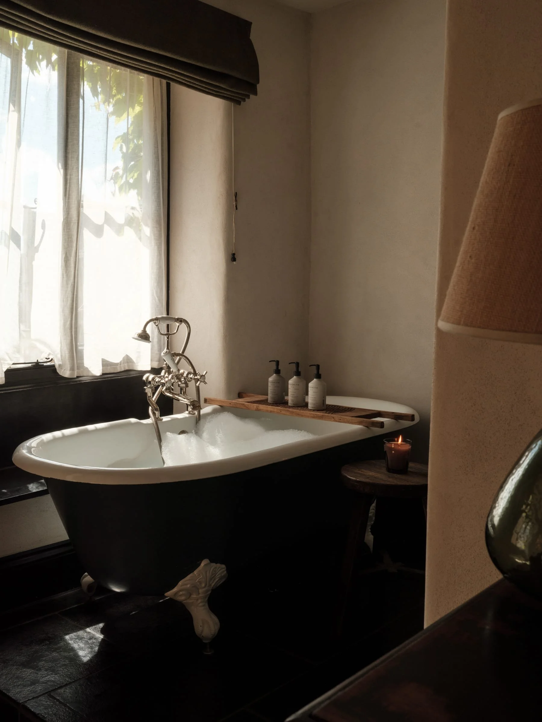 A bathtub filled with bubbles next to a window with sheer curtains, with a lit candle on a small side table, and three bottles of soap or lotion on a tray behind the bathtub.
