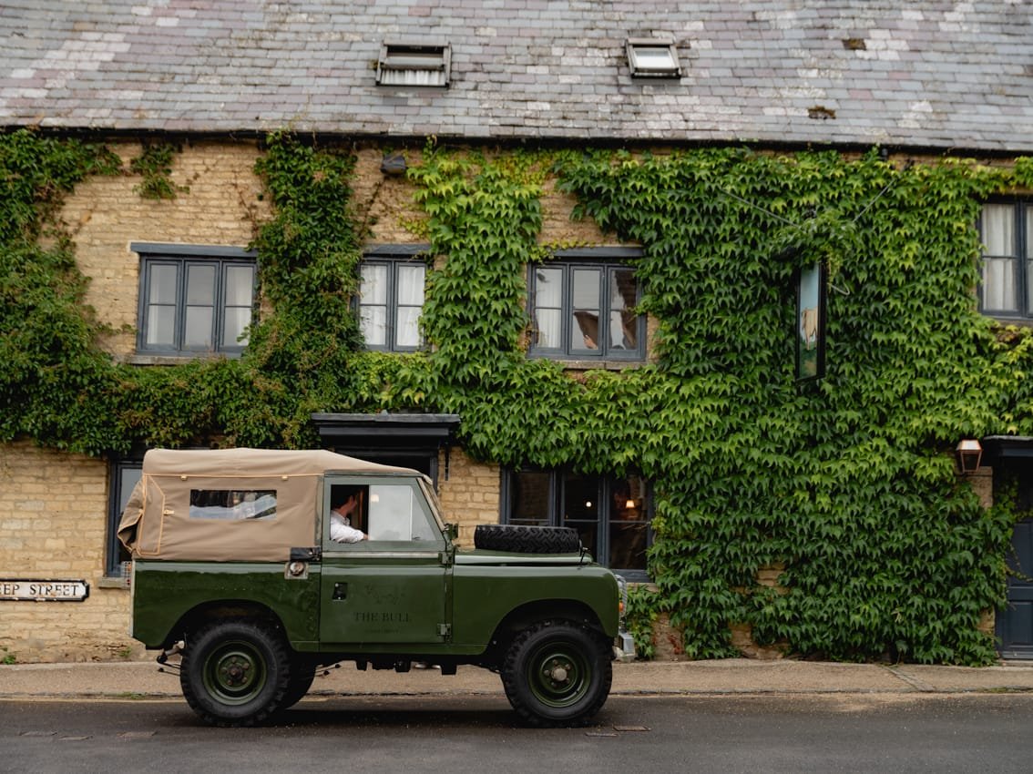 A green vintage Land Rover Defender parked on a street in front of a brick building covered in green ivy. The building has several windows and a sign reading 'Keep Street'.