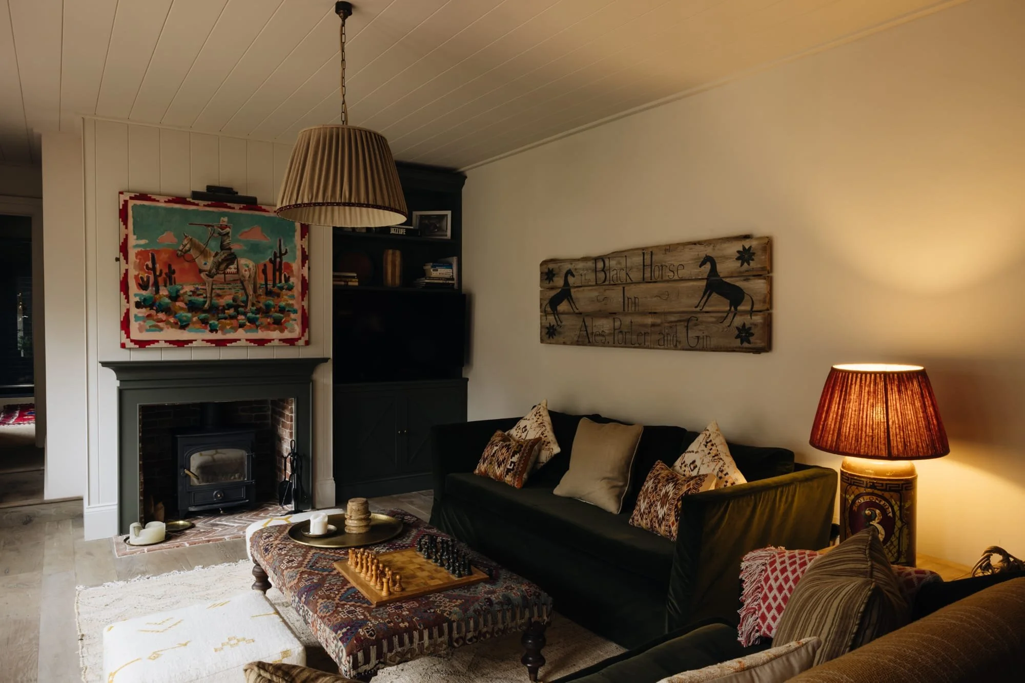 Cozy living room with a black sofa, decorative pillows, wooden coffee table with a chess set and candles, fireplace, artwork of a cowboy on horseback, wooden sign with horse images and text, and warm lighting from table lamps and ceiling fixture.