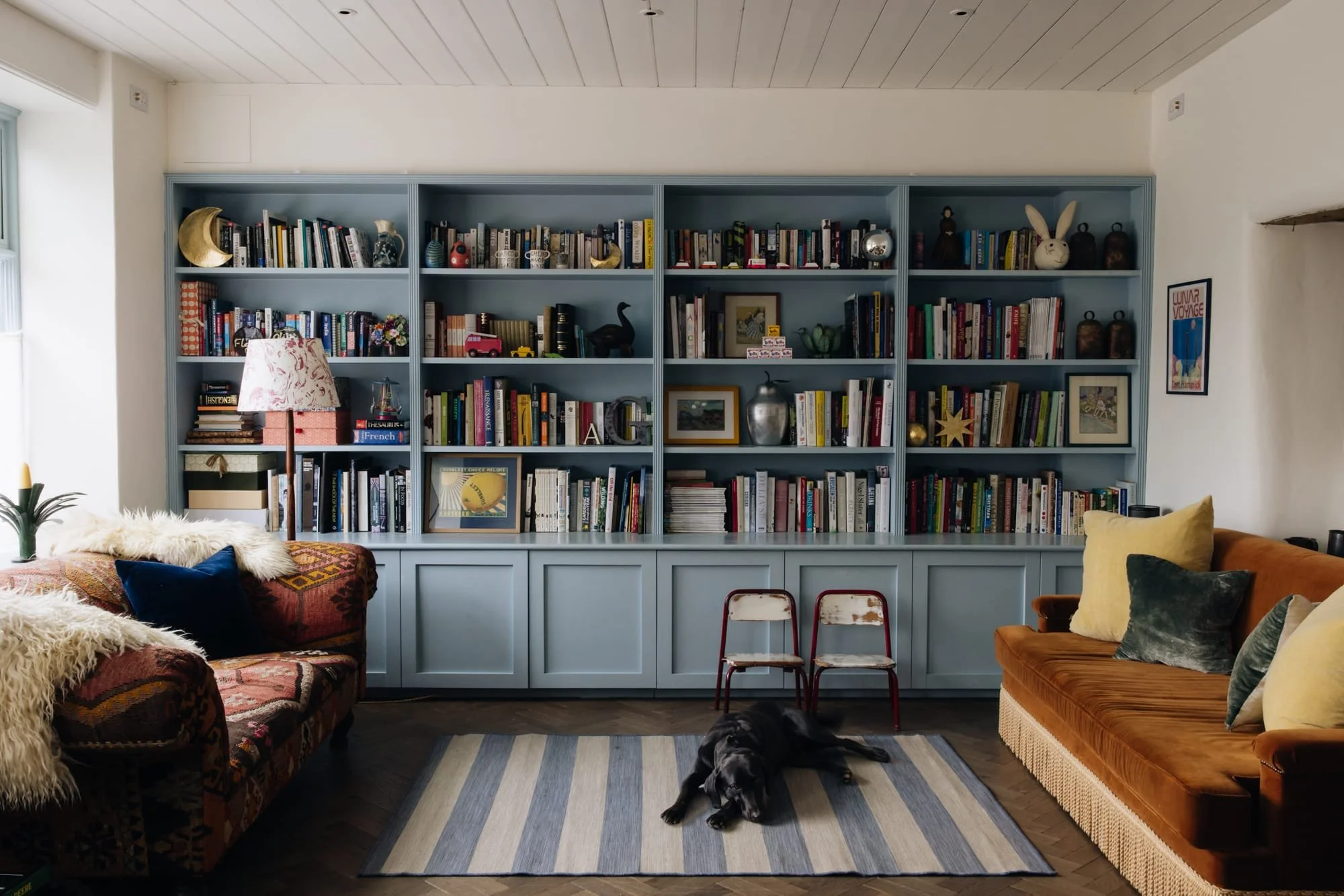 Living room with a large blue bookshelf filled with books and decorative items, two vintage red and white chairs, a brown velvet sofa with pillows, a furry throw on an armchair, a striped rug, and a black dog lying on the floor.
