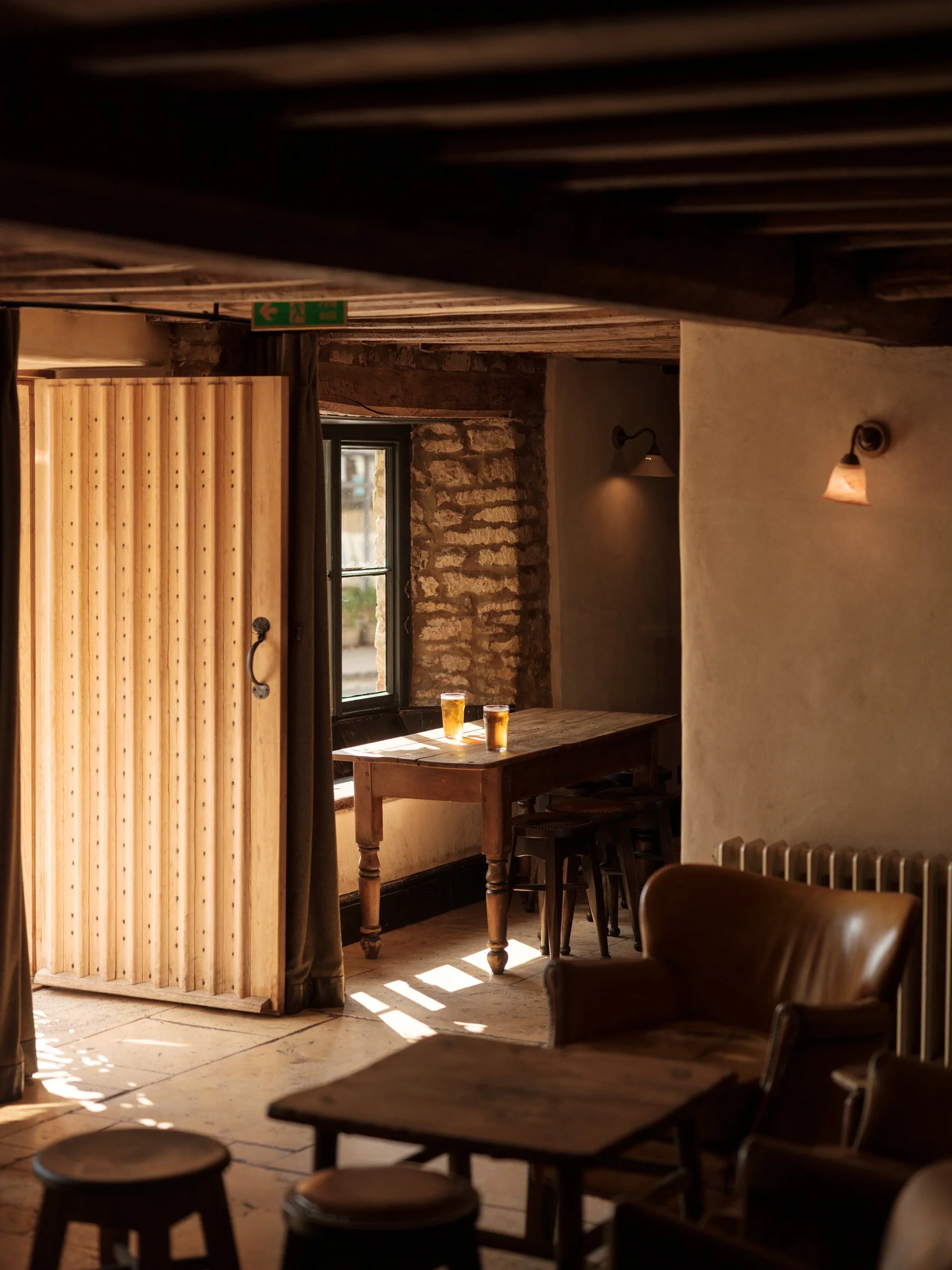 Interior of a cozy cafe or pub with wooden furniture, exposed brick wall, sunlight streaming through a window, and two glasses of beer on a table.