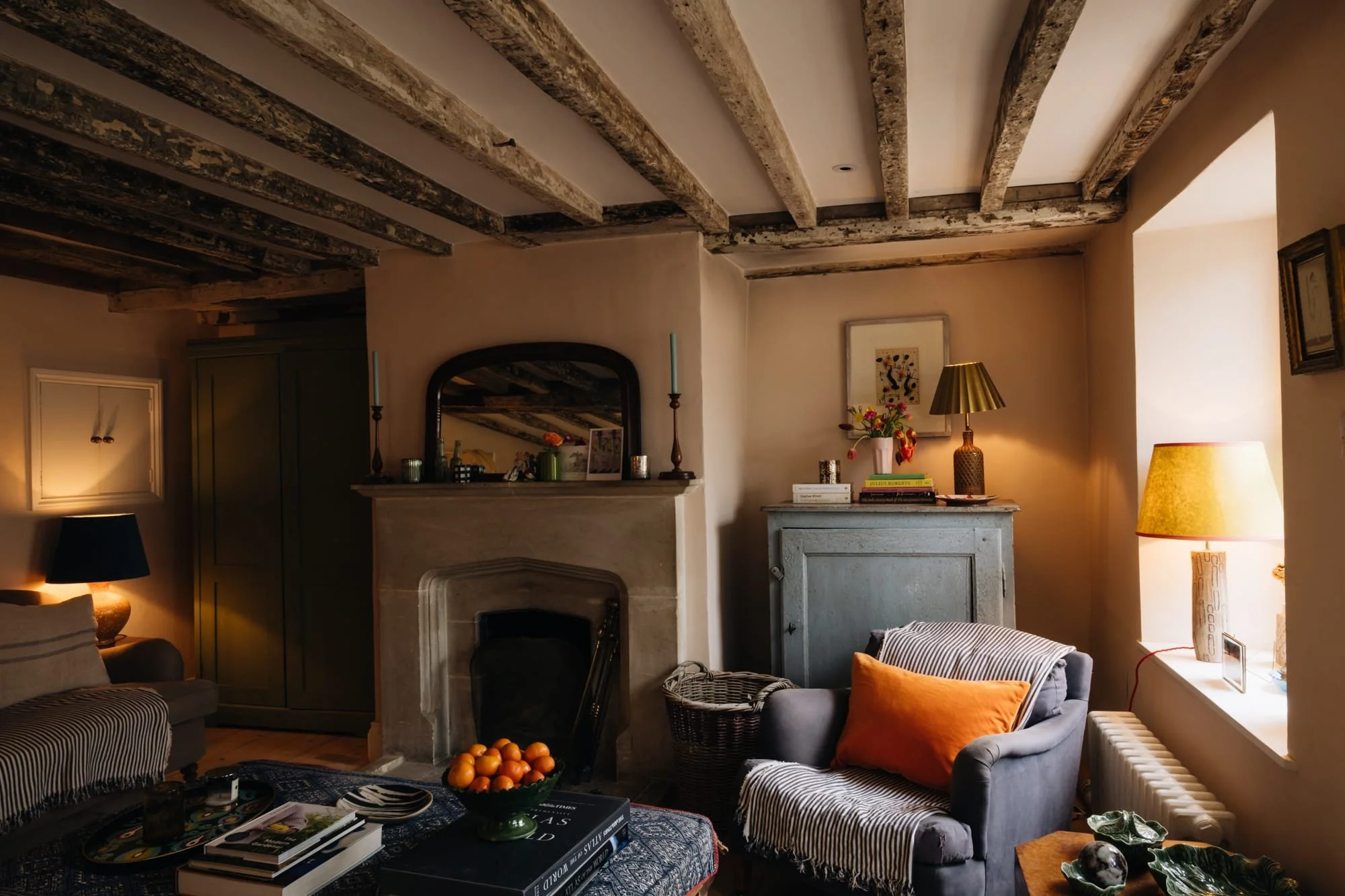 A cozy living room with rustic exposed ceiling beams, a stone fireplace, and various furniture pieces including armchairs, tables, and books. The room features warm lighting, decorative items, and artwork on the walls.