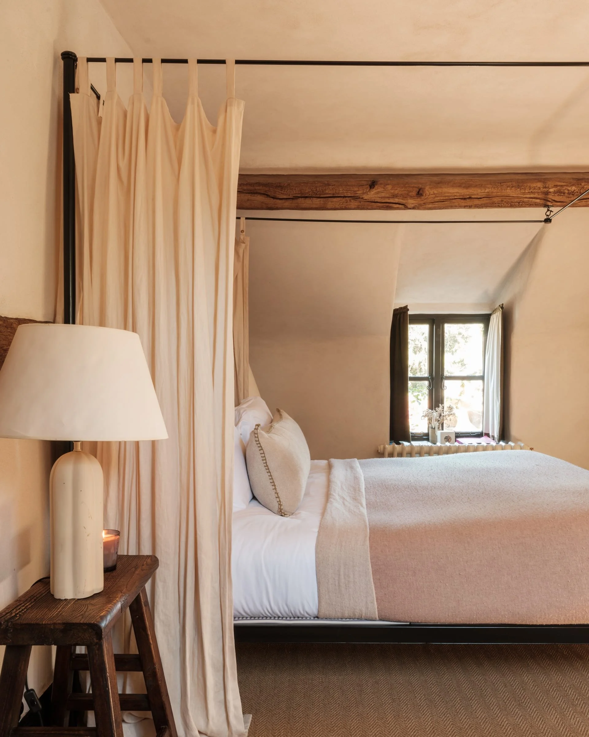 Cozy bedroom with a bed, pillows, and beige bedding, a window with curtains, a wooden side table with a lamp and a candle, and a rustic wooden beam ceiling.