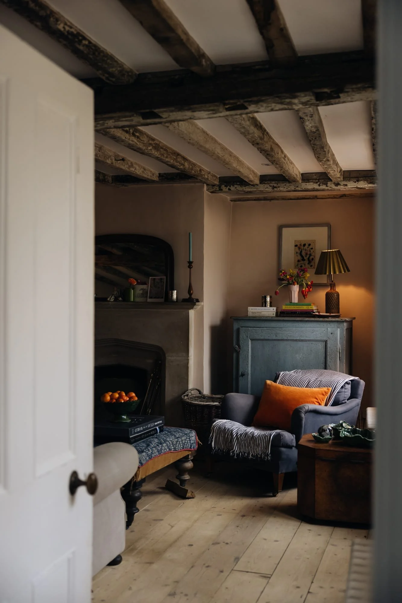 Cozy living room with wooden beams on the ceiling, a fireplace, an armchair with a cushion, a blue cabinet, and a lamp, illuminated by warm lighting.