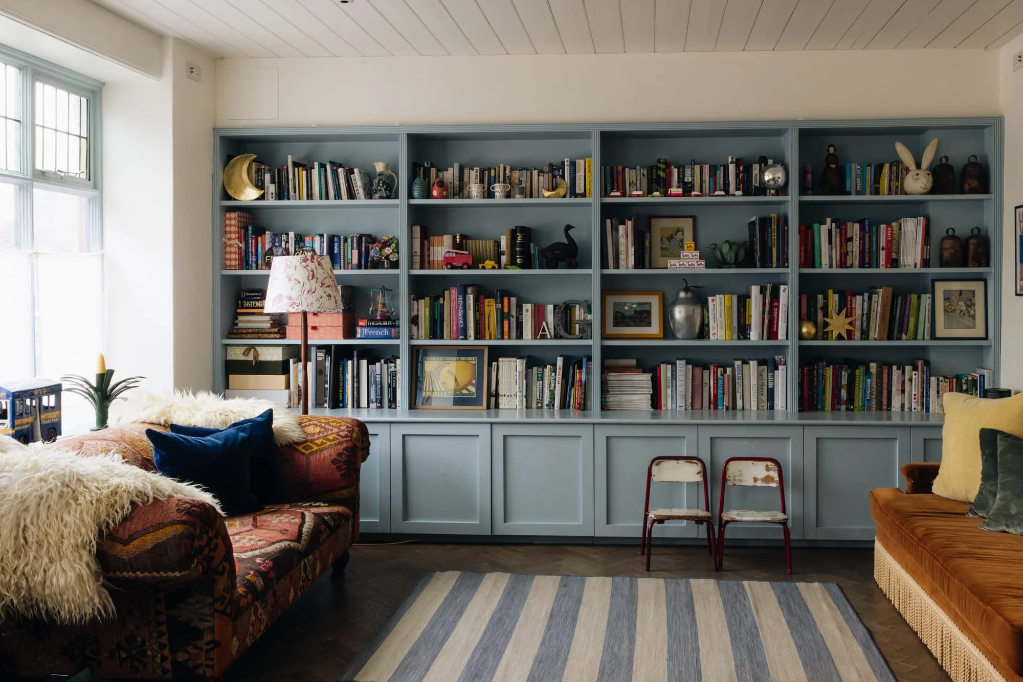 Living room with a large blue bookshelf filled with books, decorations, and framed photos. Two couches, one with a colorful pattern and fluffy throws, the other plain brown, are arranged with pillows. Between the couches are two small vintage chairs.