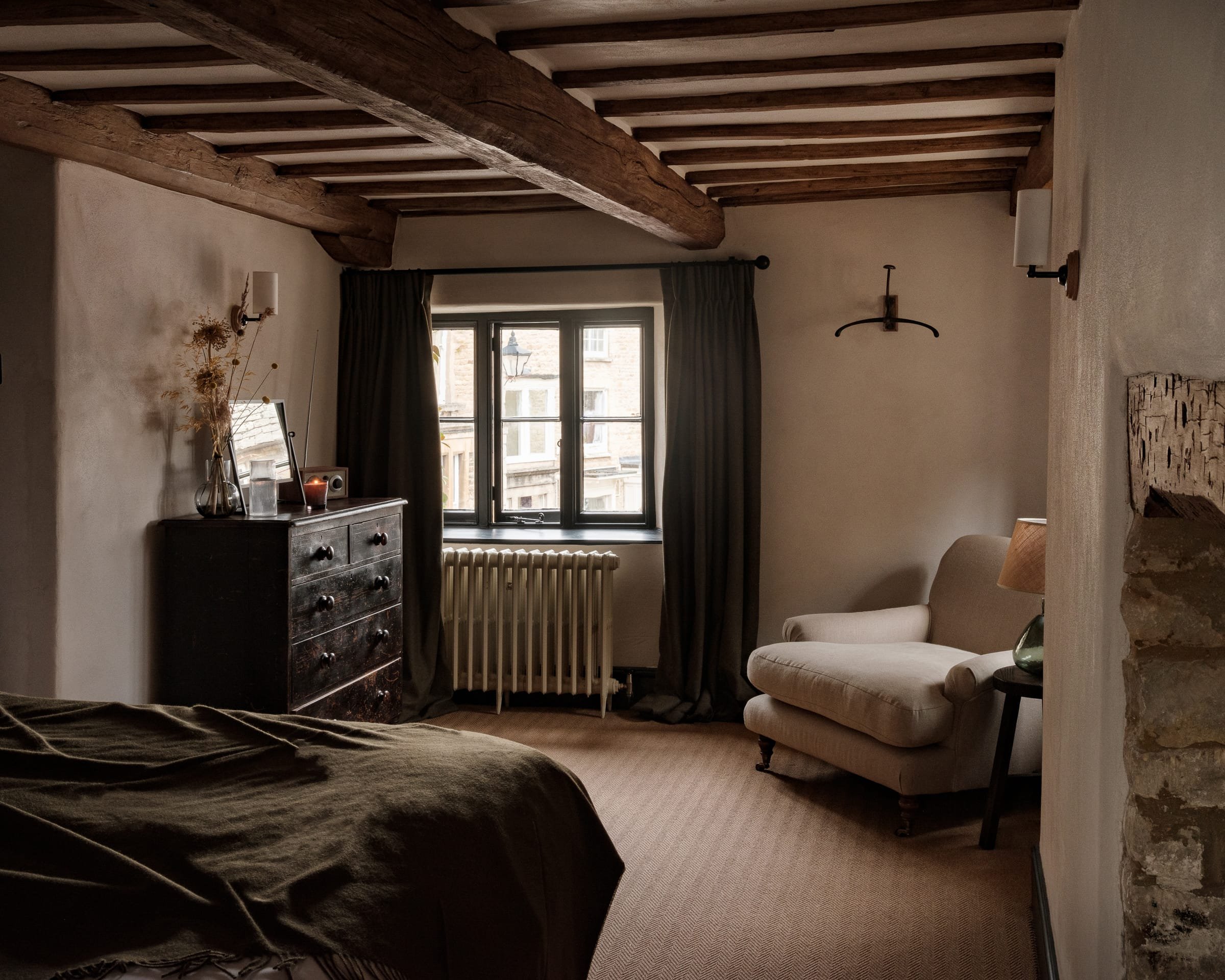 A cozy bedroom with wooden ceiling beams, a window with dark curtains, a beige armchair with a side table and lamp, a black dresser with decorative items, and a bed with a dark blanket.
