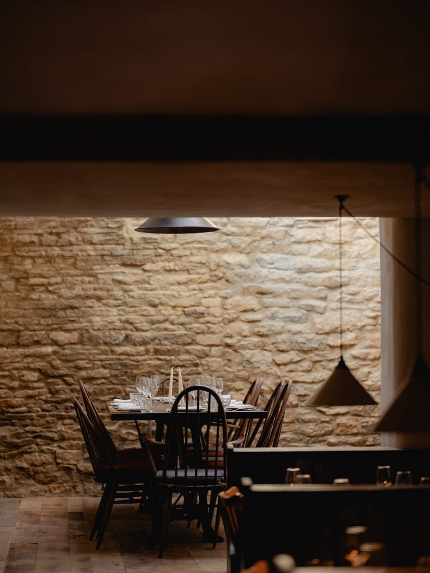 A cozy dining room with a brick wall, a table set with wine glasses, and wooden chairs, illuminated by pendant and ceiling lights.