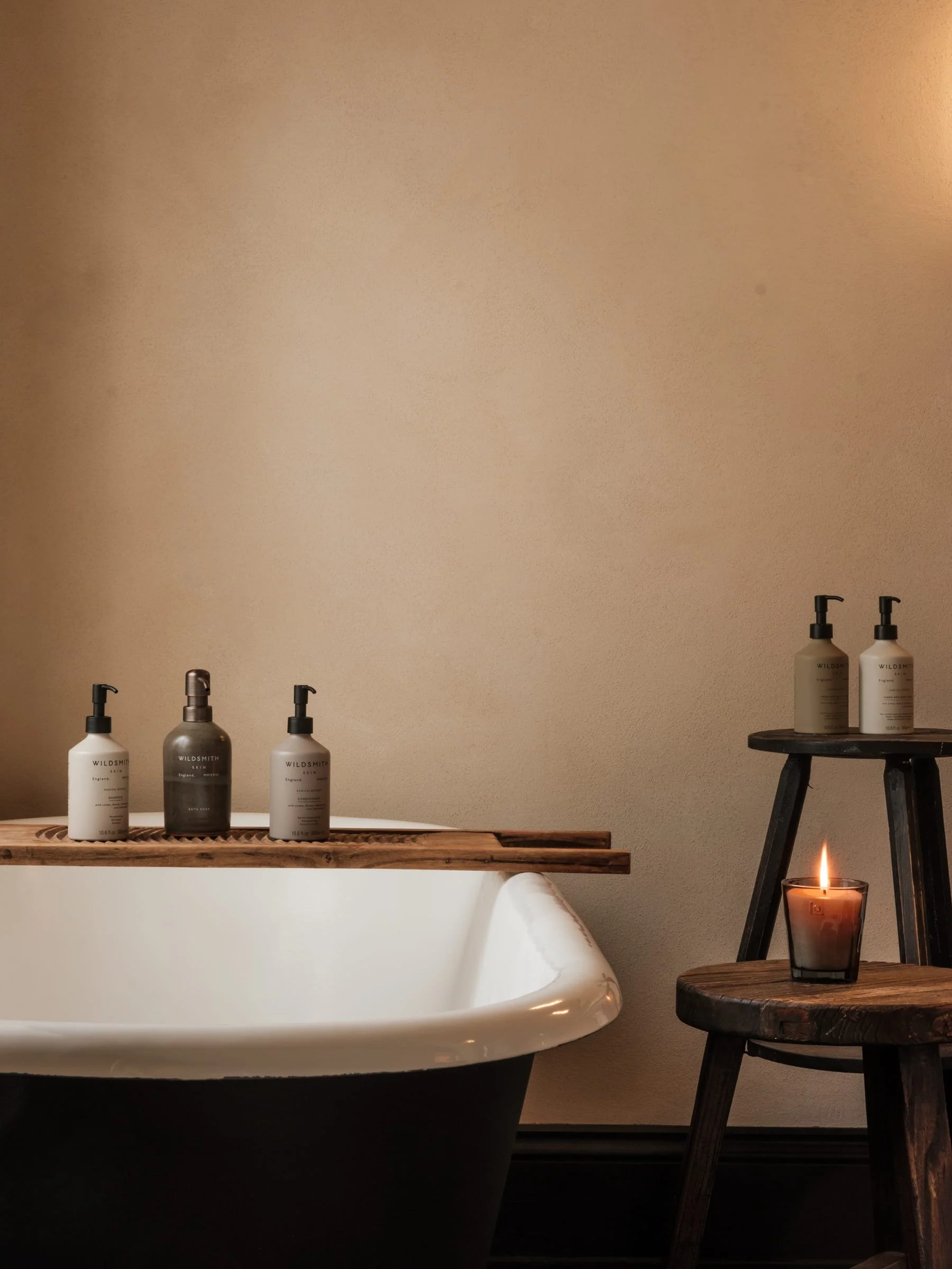 A bathroom scene with a white bathtub, three bottles of Wildsmith skincare products on a wooden tray on the edge of the tub, and two more bottles on a wooden stool. A lit candle is on a small wooden table next to the stool, creating a cozy atmosphere