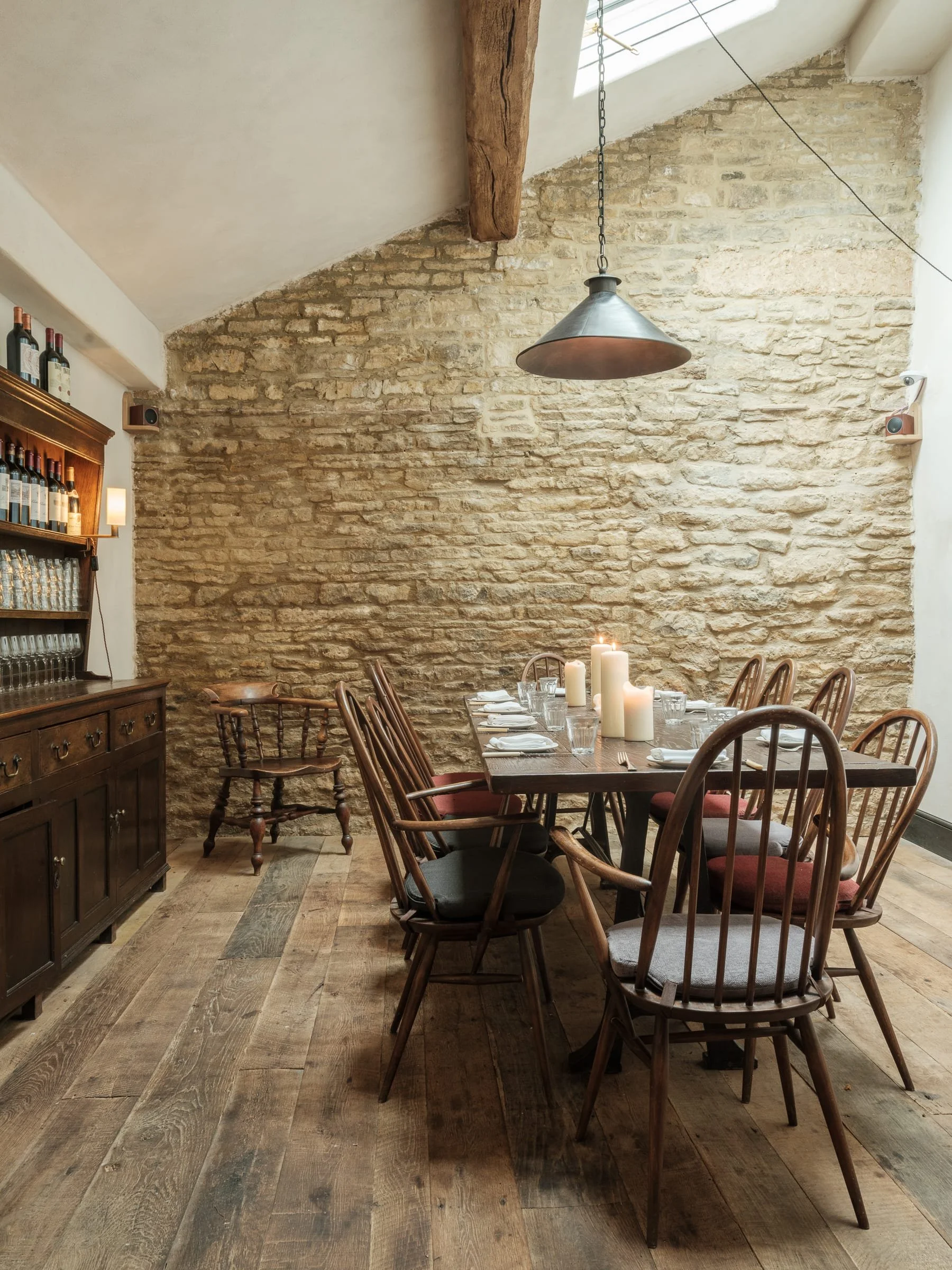 Interior of a dining room with a long wooden table set with candles, glasses, and plates, surrounded by wooden chairs, against a stone wall with a skylight overhead.