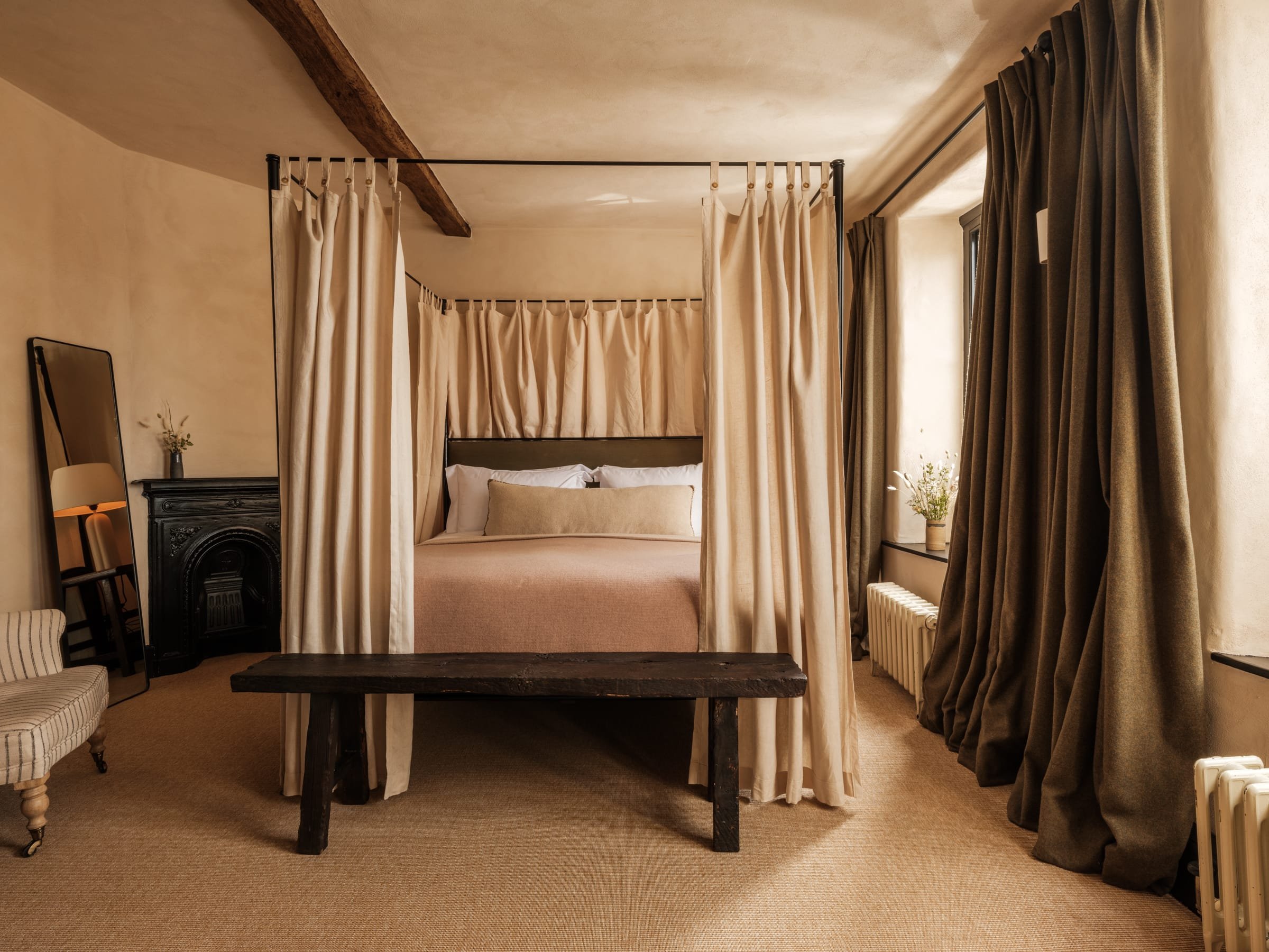 A bedroom with a four-poster bed with cream-colored curtains. There are dark curtains on the windows, a vintage black fireplace, a large mirror, a bench at the foot of the bed, and small vases of flowers on the windowsills. The room has beige walls a