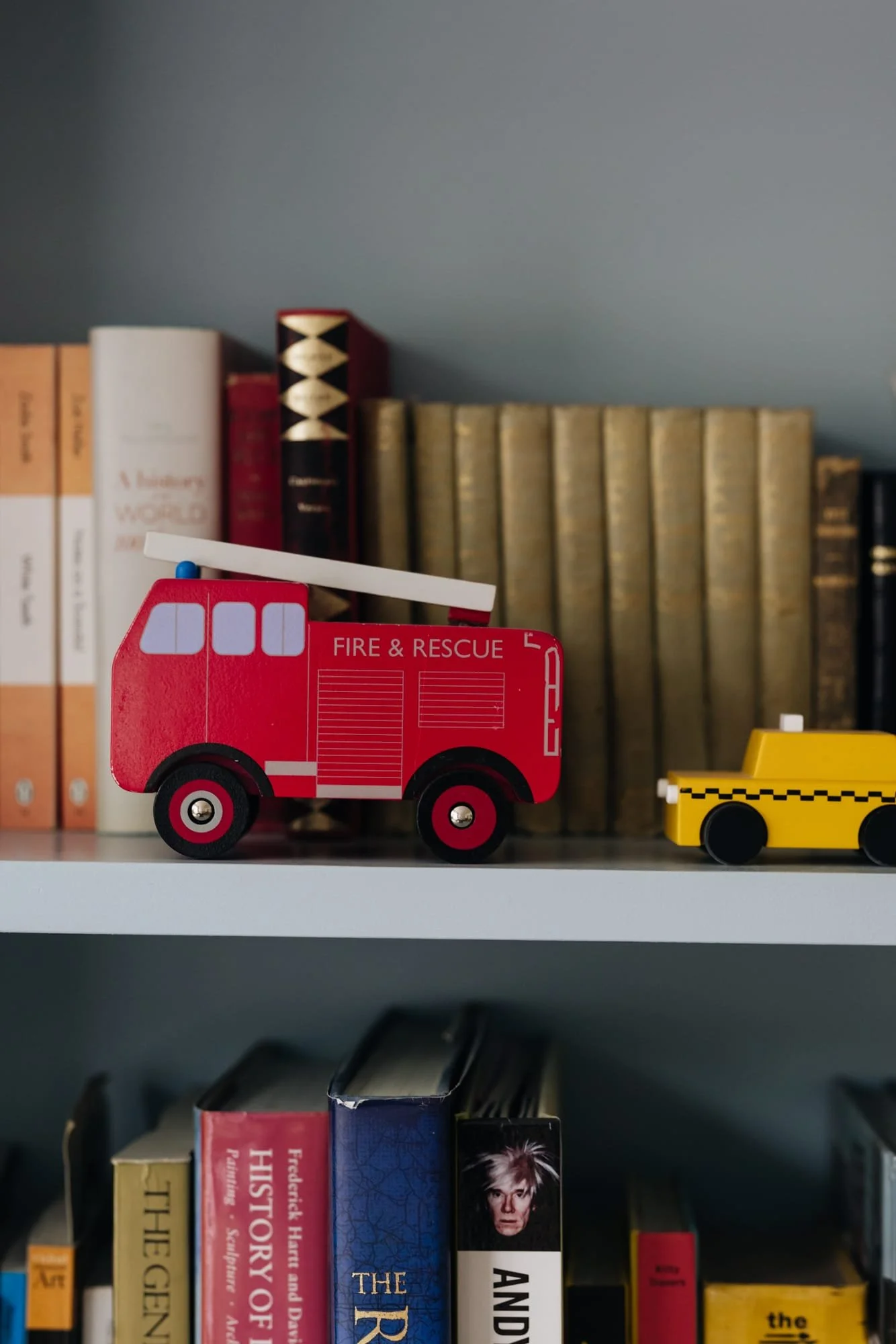 Toy fire truck and toy yellow taxi on a bookshelf with books in the background.
