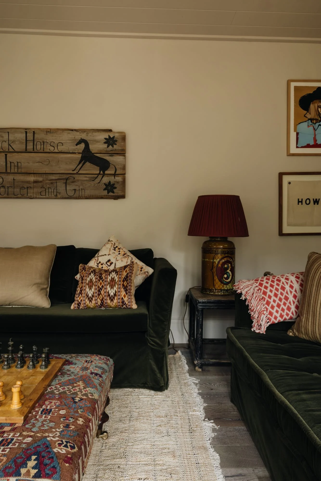 Living room with two black sofas, various decorative pillows, a wooden chessboard on a patterned rug, a vintage lamp with a red shade, and wall art including a 