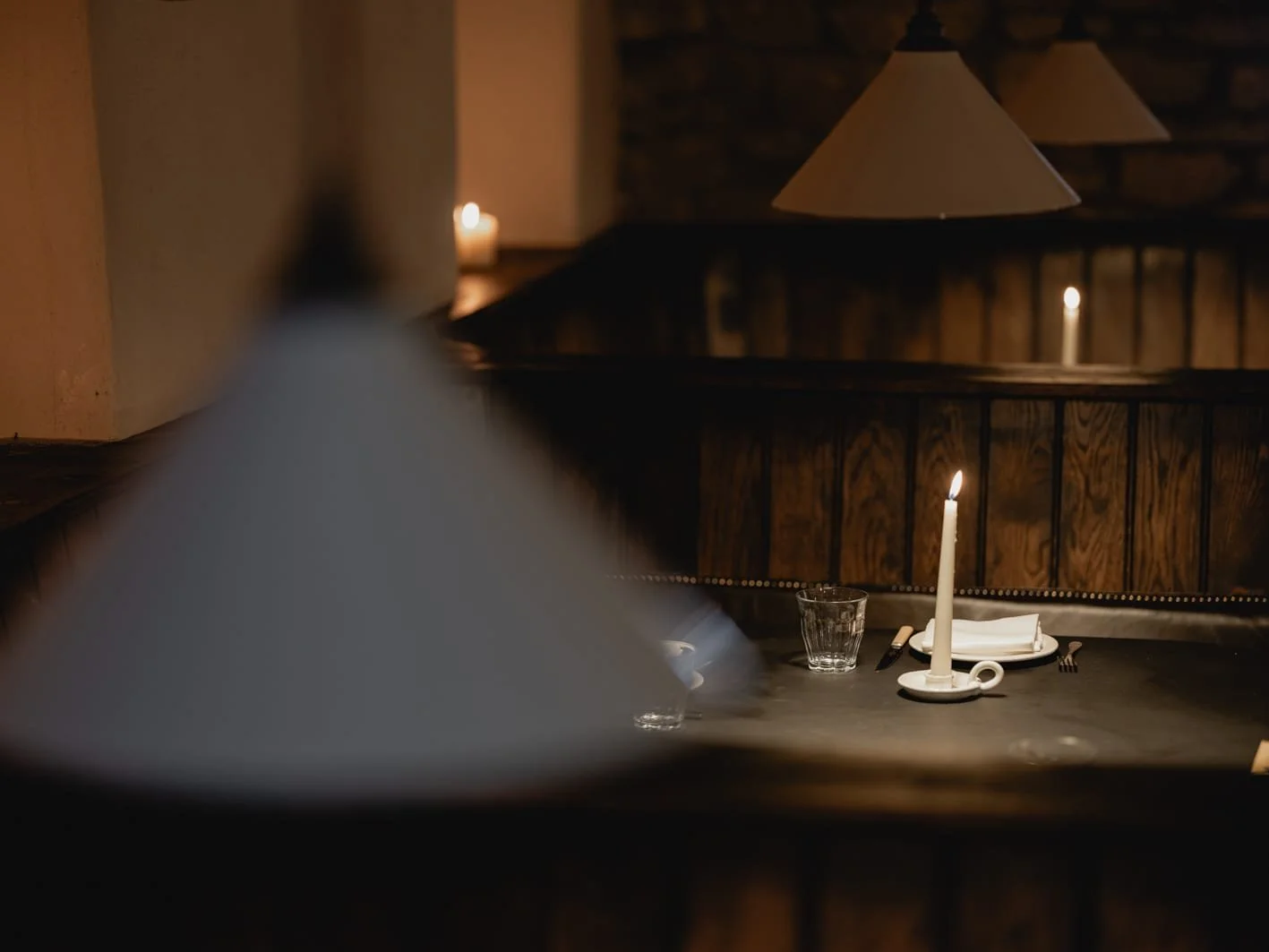 A dimly lit restaurant table set with a lit candle, a glass, a plate with a napkin, and cutlery, with hanging lamps and a dark wooden background.