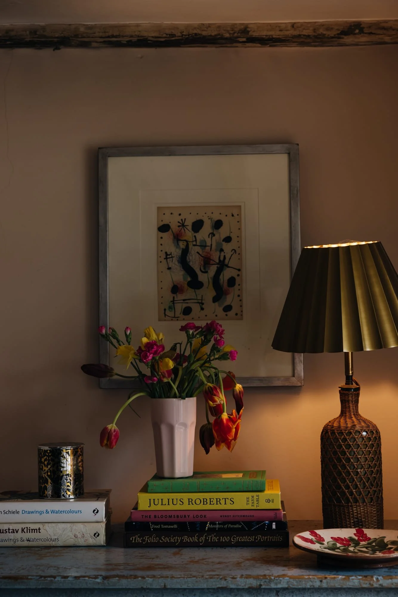 A wooden table with a stack of books, a white vase with pink and yellow flowers, a patterned container, a decorative plate, a table lamp with a gold shade, a framed abstract artwork on the wall behind.