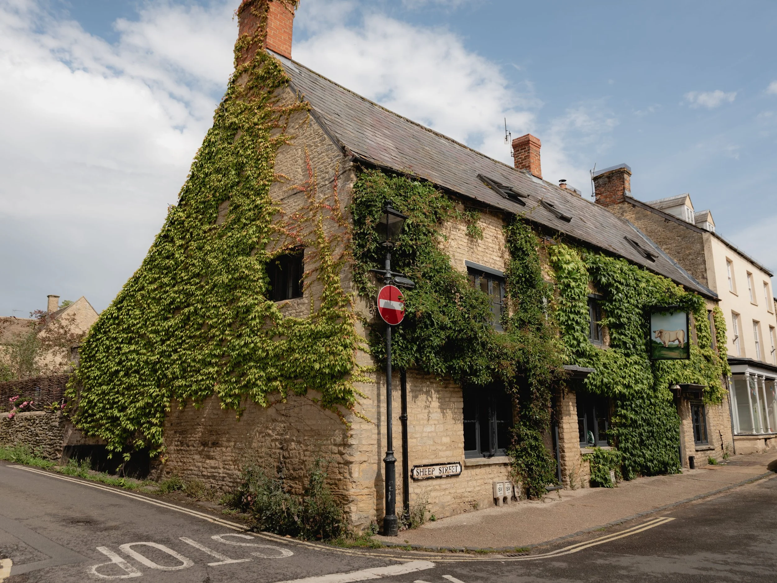 A stone house with a sloped roof covered in green ivy vines, located at a street corner with a street sign reading 'Sheep Street' and a no entry traffic sign. The house has multiple small windows and a sign depicting a sheep. The scene is set on a pa