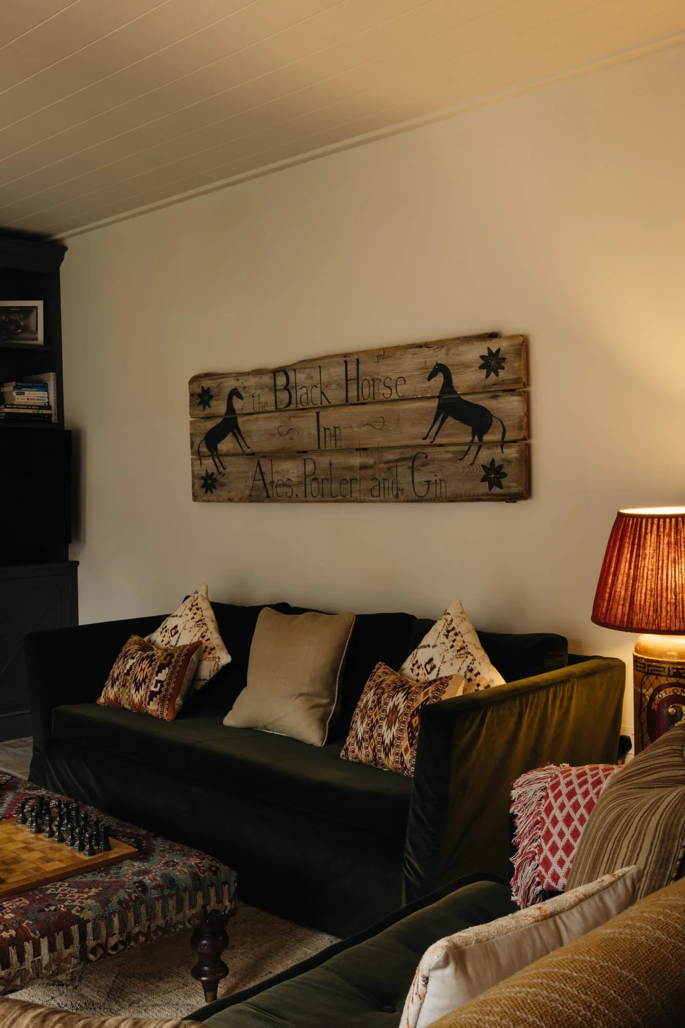 Living room with a black sofa, decorative pillows, a wooden sign on the wall reading 'The Black Horse Inn Ales, Porter and Gin' with horse illustrations, a table with a chess set, and a lamp with a red shade.