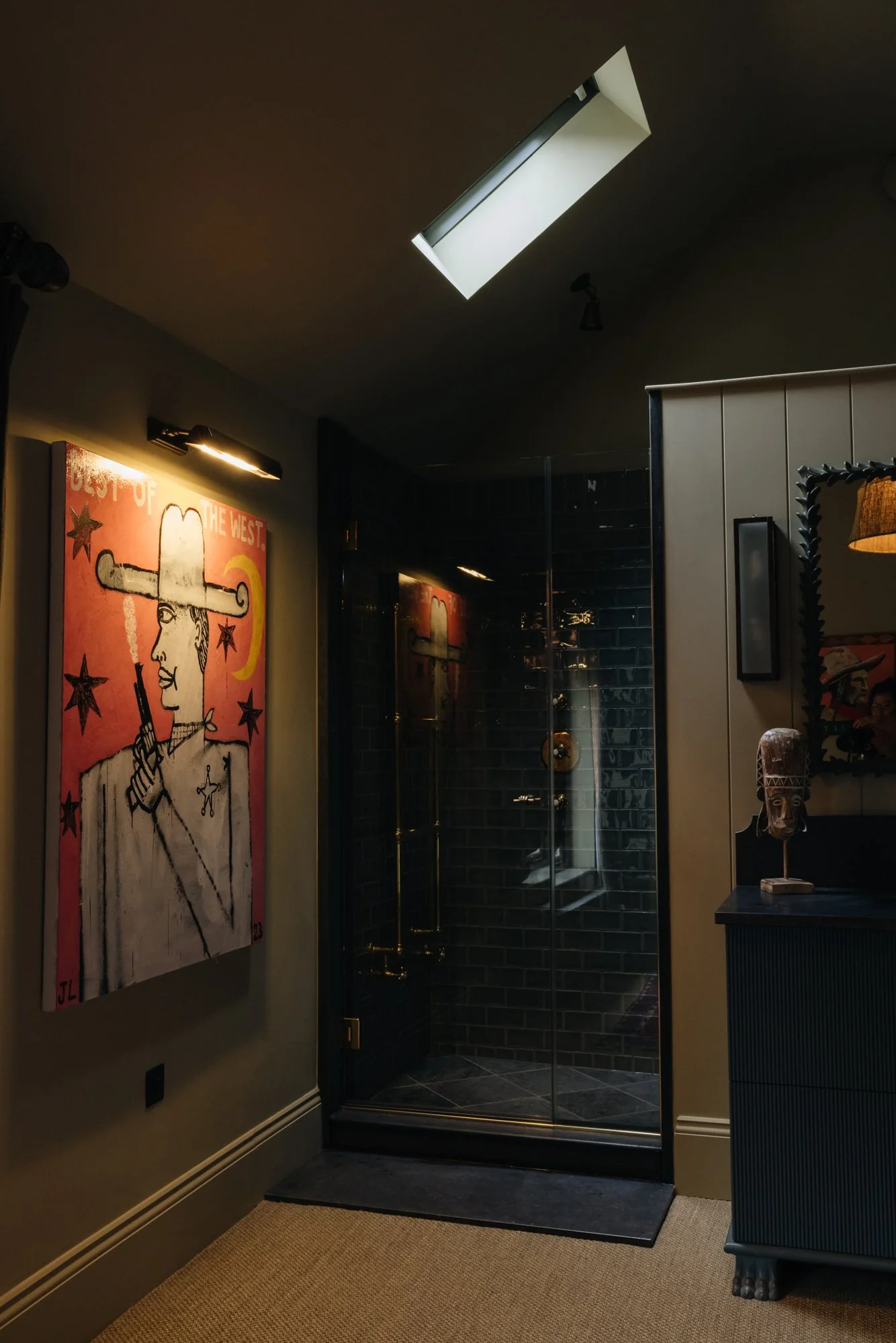 Modern bathroom with a glass shower enclosure, black tiled walls, and a colorful large artwork on the beige wall showing a stylized figure with a hat, stars, and the text "Best of the West."
