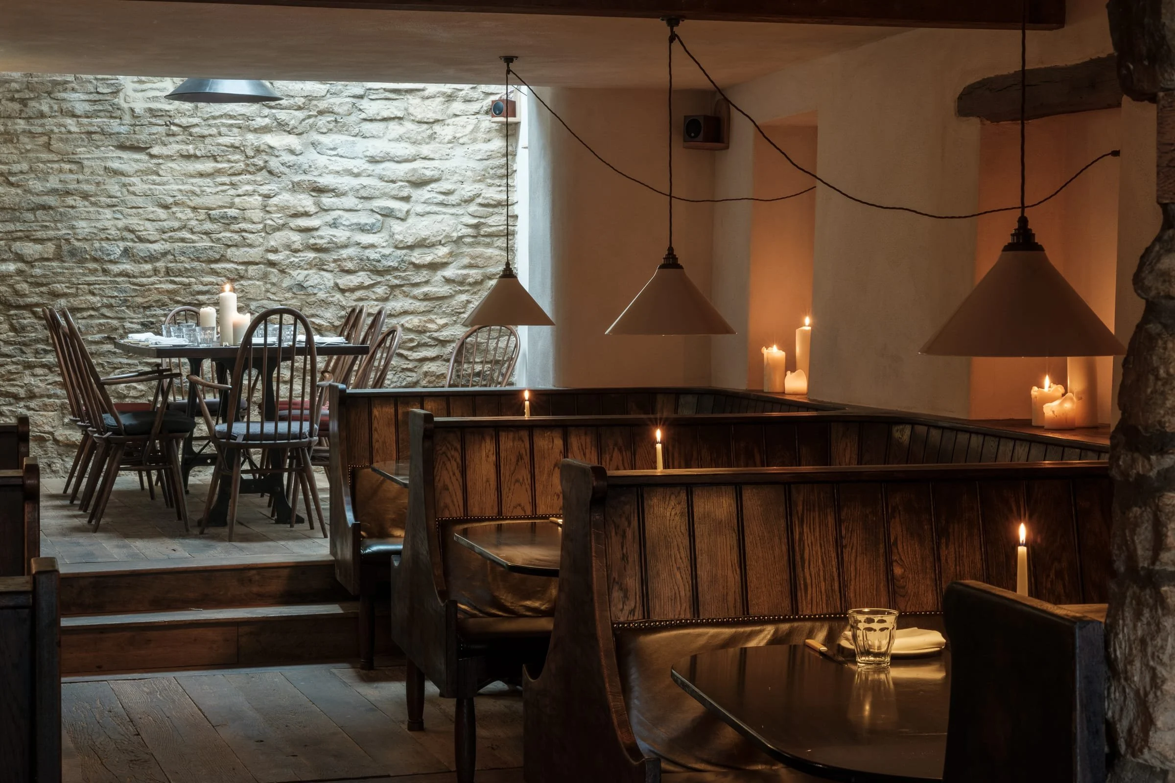 Cozy restaurant interior with wooden booths and dining tables, lit by candles and pendant lights, stone wall backdrop and rustic decor.