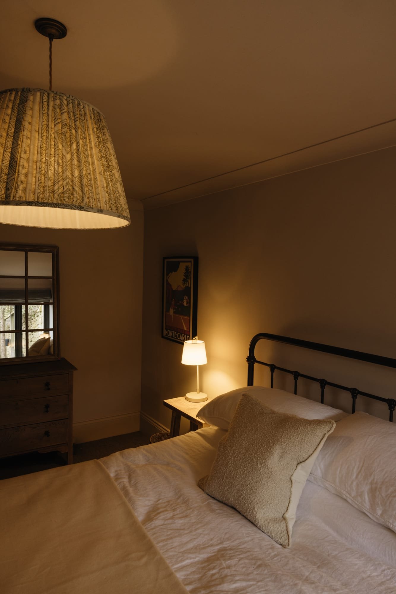 A cozy bedroom with a bed, a nightstand with a lamp, a window, and a ceiling light fixture.