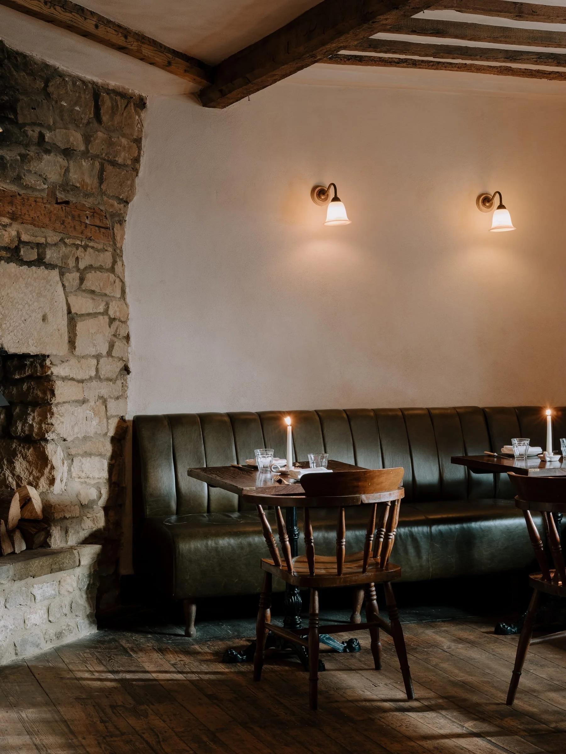 Cozy restaurant corner with lit candles on tables, wooden chairs, a green banquette, stone fireplace on the left, and wall-mounted lights on a white wall.