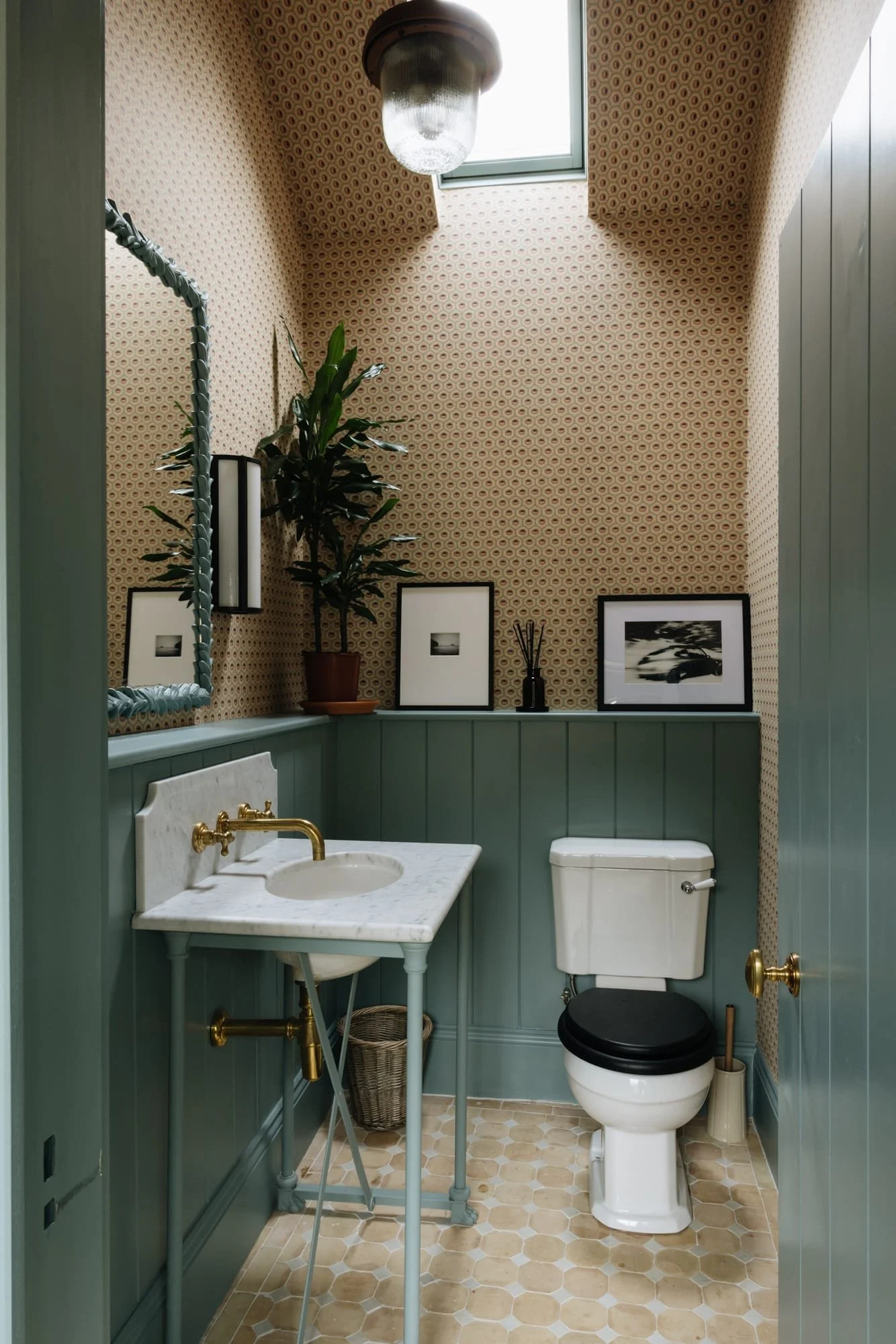 A small vintage-style bathroom with patterned wallpaper, green wainscoting, a small marble sink, a toilet with a black seat, and decorative framed black-and-white artwork and a plant on the shelf. There is a skylight above and an overhead light fixtu