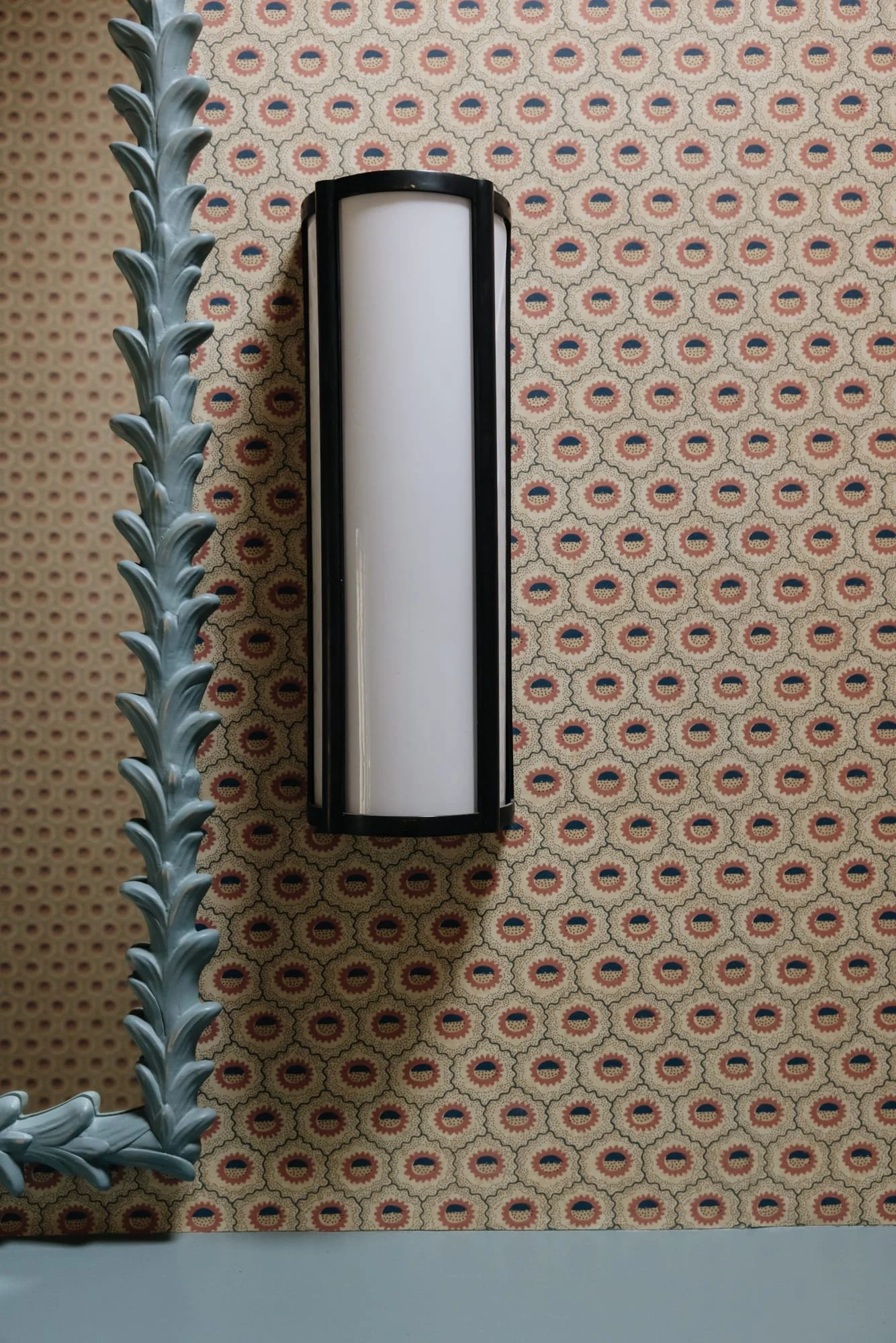 Wall-mounted light fixture with a white frosted glass cover, a decorative mirror with an ornate blue frame, and patterned wallpaper with red, blue, and beige geometric designs.