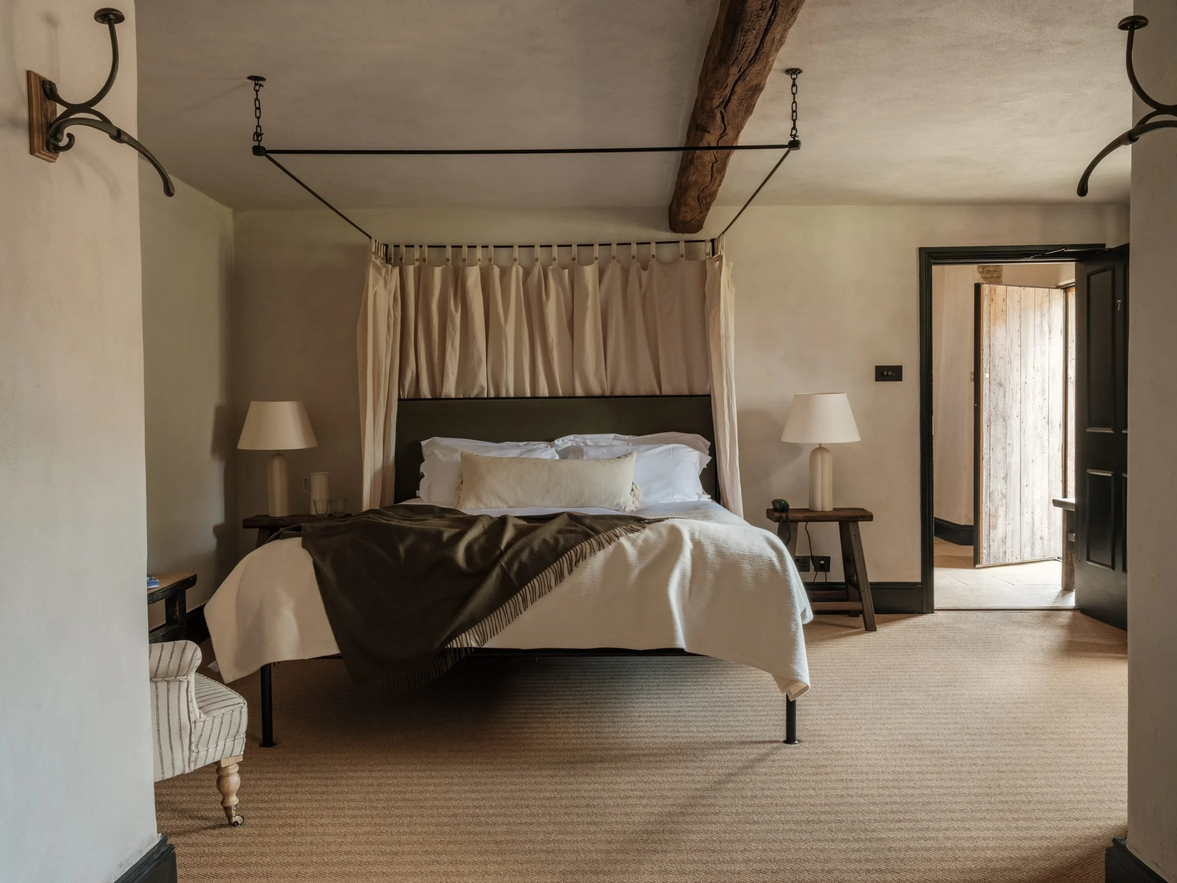 A cozy bedroom with a bed centered against the wall, topped with white bedding and a brown throw blanket. Flanking the bed are two matching bedside tables with white lamps. The room features a rustic aesthetic with a wooden beam on the ceiling and a 