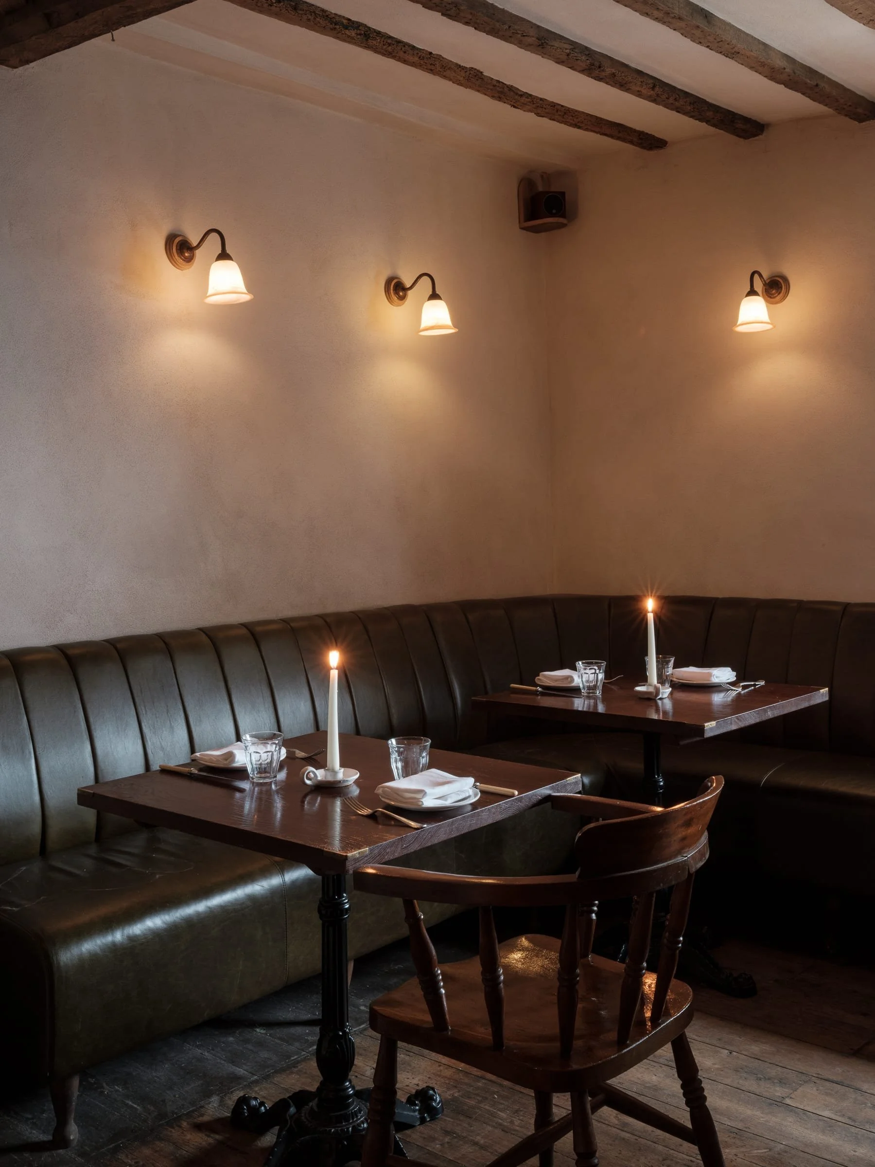 A cozy restaurant corner with two dark wooden tables set with white napkins, glasses, and lit candles. There are two wooden chairs and a curved leather banquette against a beige wall with three wall lamps, creating an intimate ambiance.
