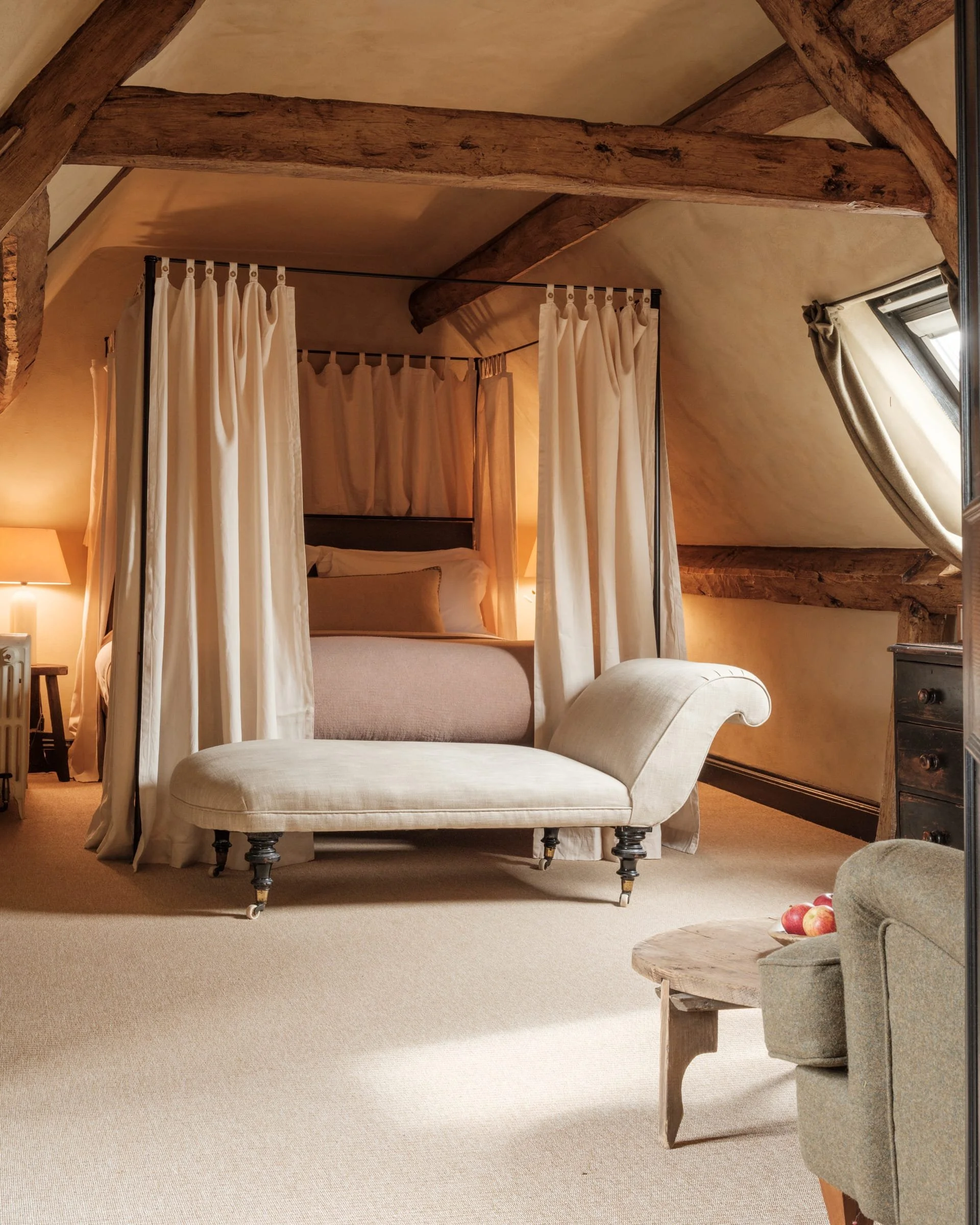Elegant bedroom with a four-poster canopy bed with cream curtains, beige bedding, a chaise lounge at the foot of the bed, a small wooden side table with apples, a beige armchair, a rustic dresser, and a skylight window with curtains, illuminated by w
