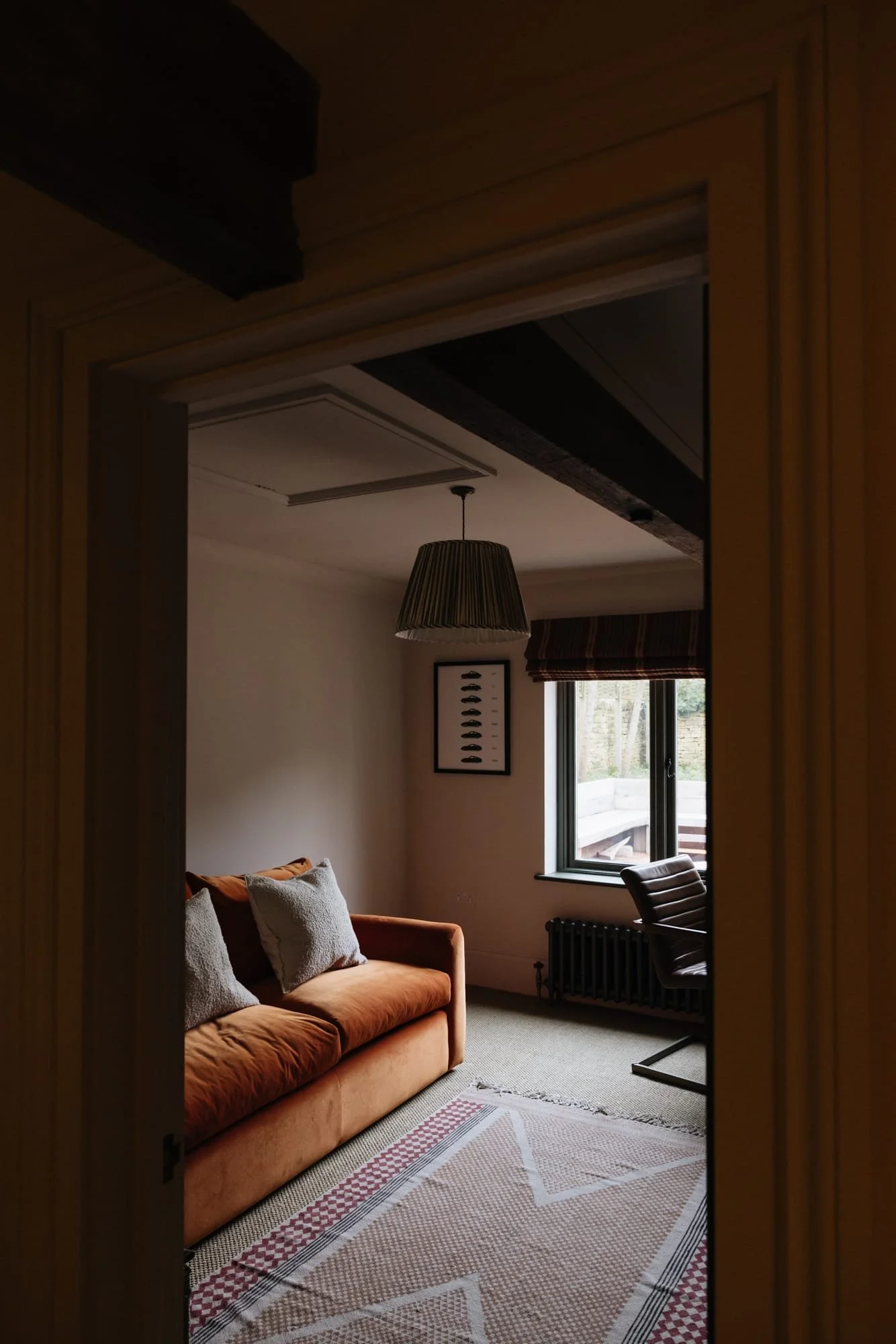 A cozy living room with a peach-colored sofa, four textured pillows, a window with a striped Roman shade, a framed art piece of fish, a beige and red patterned rug, a black desk chair, and a hanging lampshade.