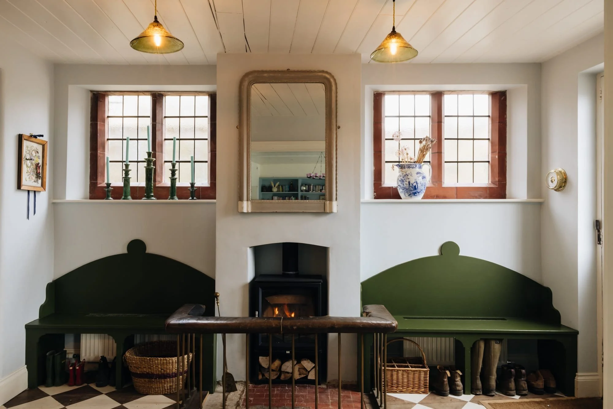 Interior of a cozy living room with two green built-in benches, a small fireplace with a fire, and a large mirror above the fireplace. There are two windows on either side of the mirror, decorated with blue and white vases and candles. The ceiling ha