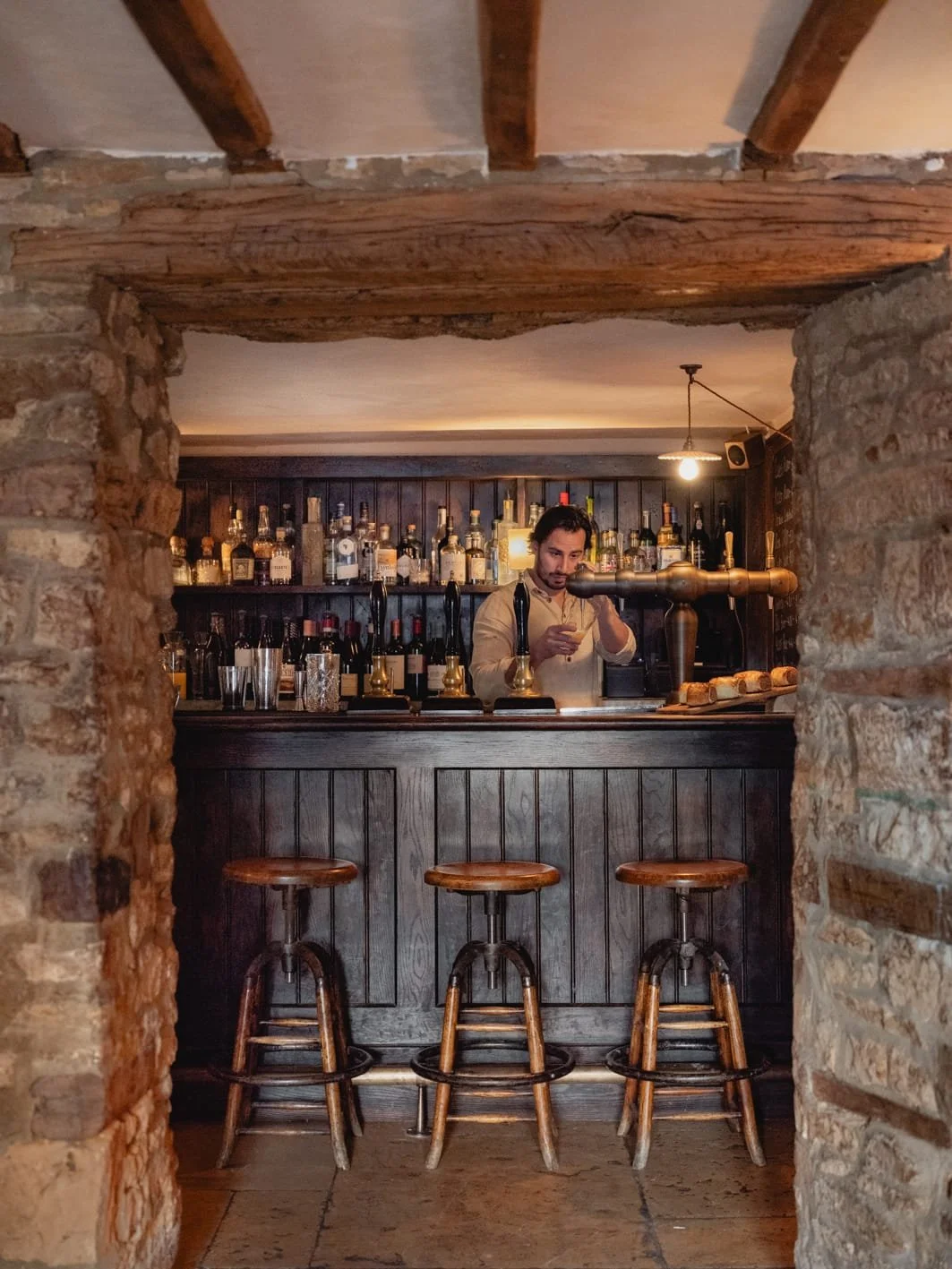 A bartender is behind a dark wooden bar preparing a drink in a cozy, rustic setting with exposed brick and wooden beams, with bottles of liquor displayed on shelves behind him.